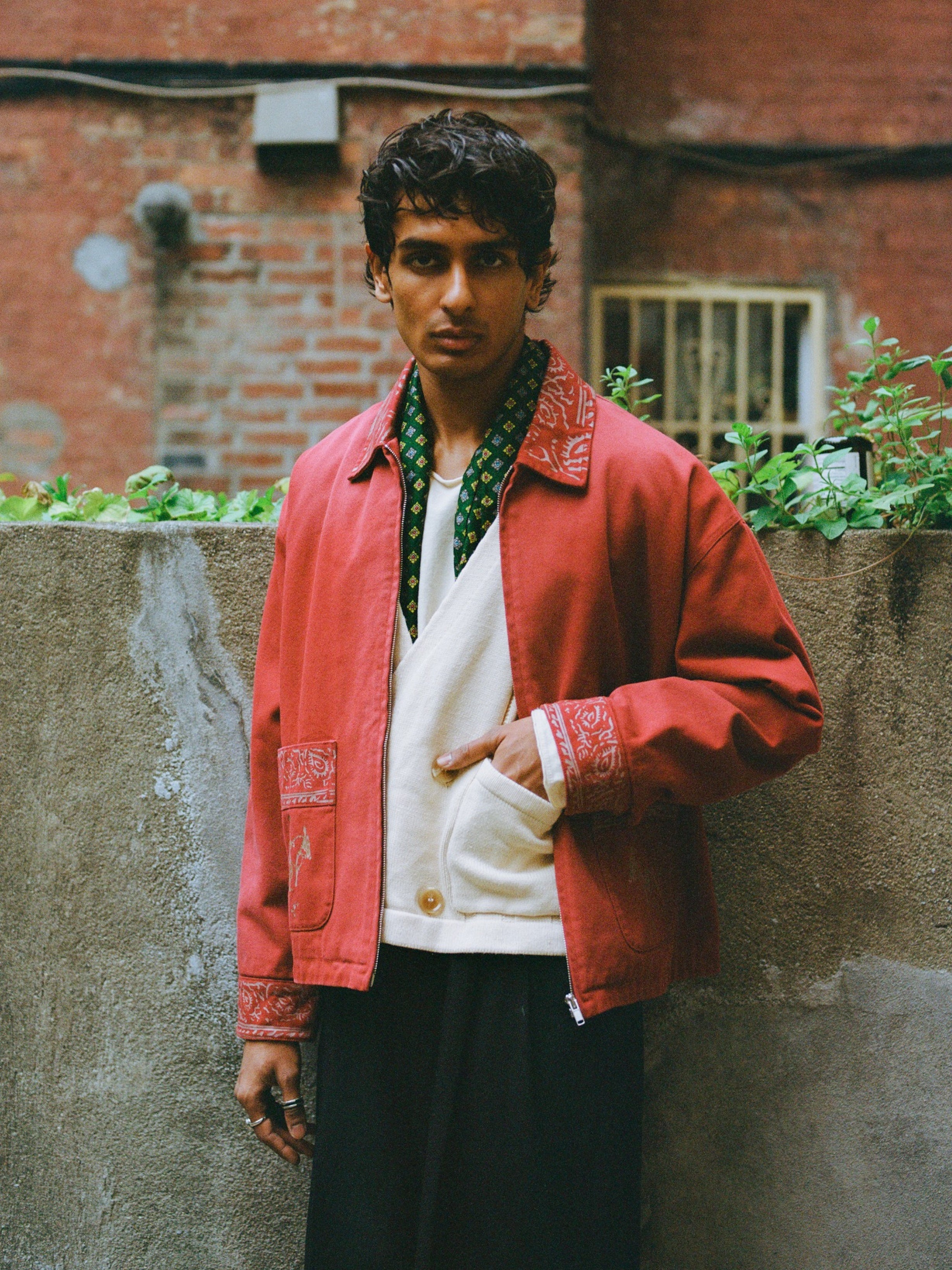 A person stands outdoors by a concrete wall, wearing Found’s Abhala Printed Work Jacket with equestrian motifs, layered over a white shirt and black pants. The unisex jacket offers a relaxed fit, and the individual has a neutral expression.