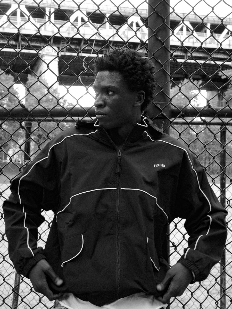A person wears the FOUND Arena Stripe Track Jacket, a unisex lightweight technical piece, standing before a chain-link fence with a bridge in the background.