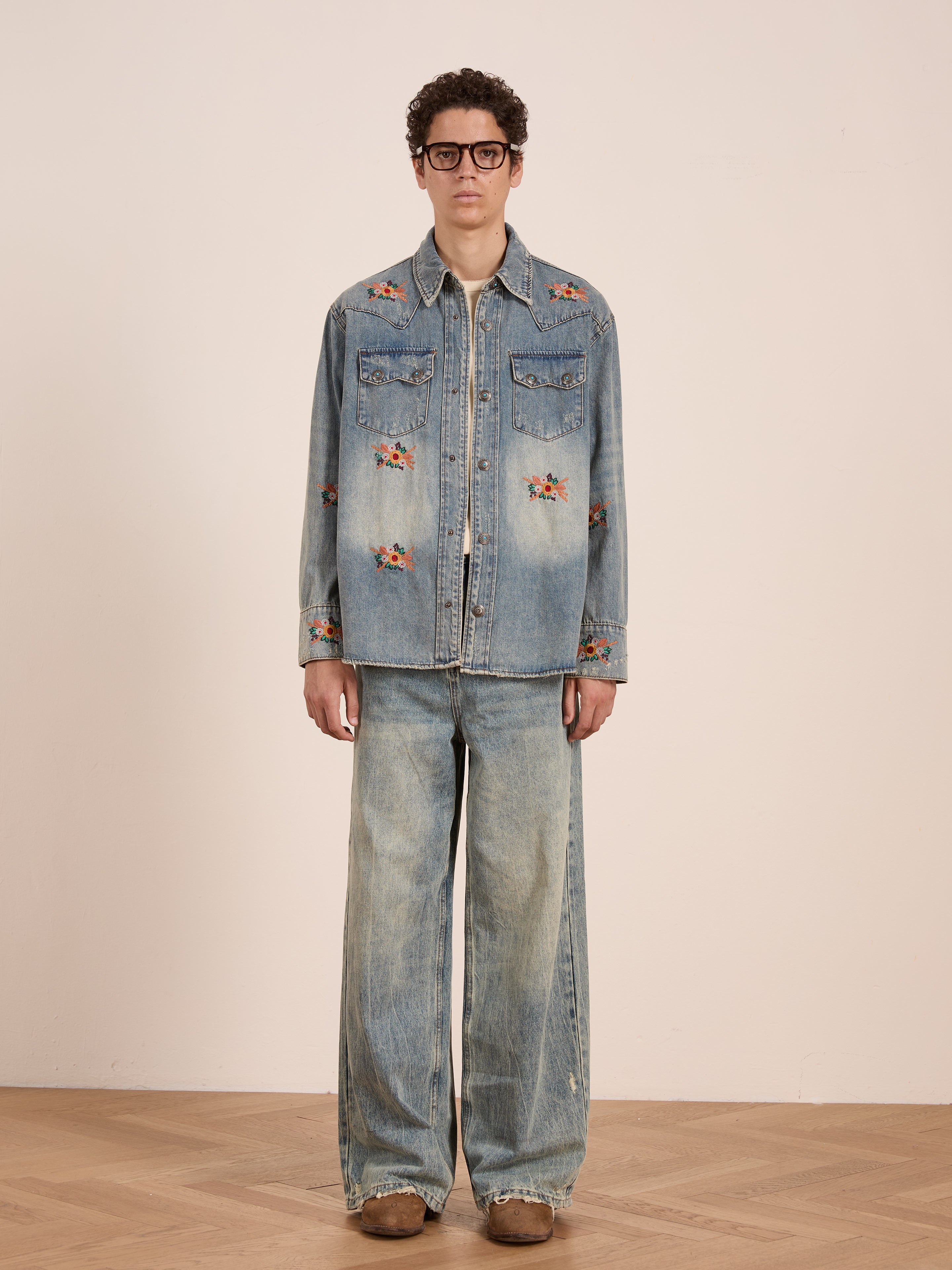 A person stands indoors in a FOUND Bouquet Floral Embroidered Western Denim Shirt, paired with matching wide-leg jeans, brown shoes, and large glasses.