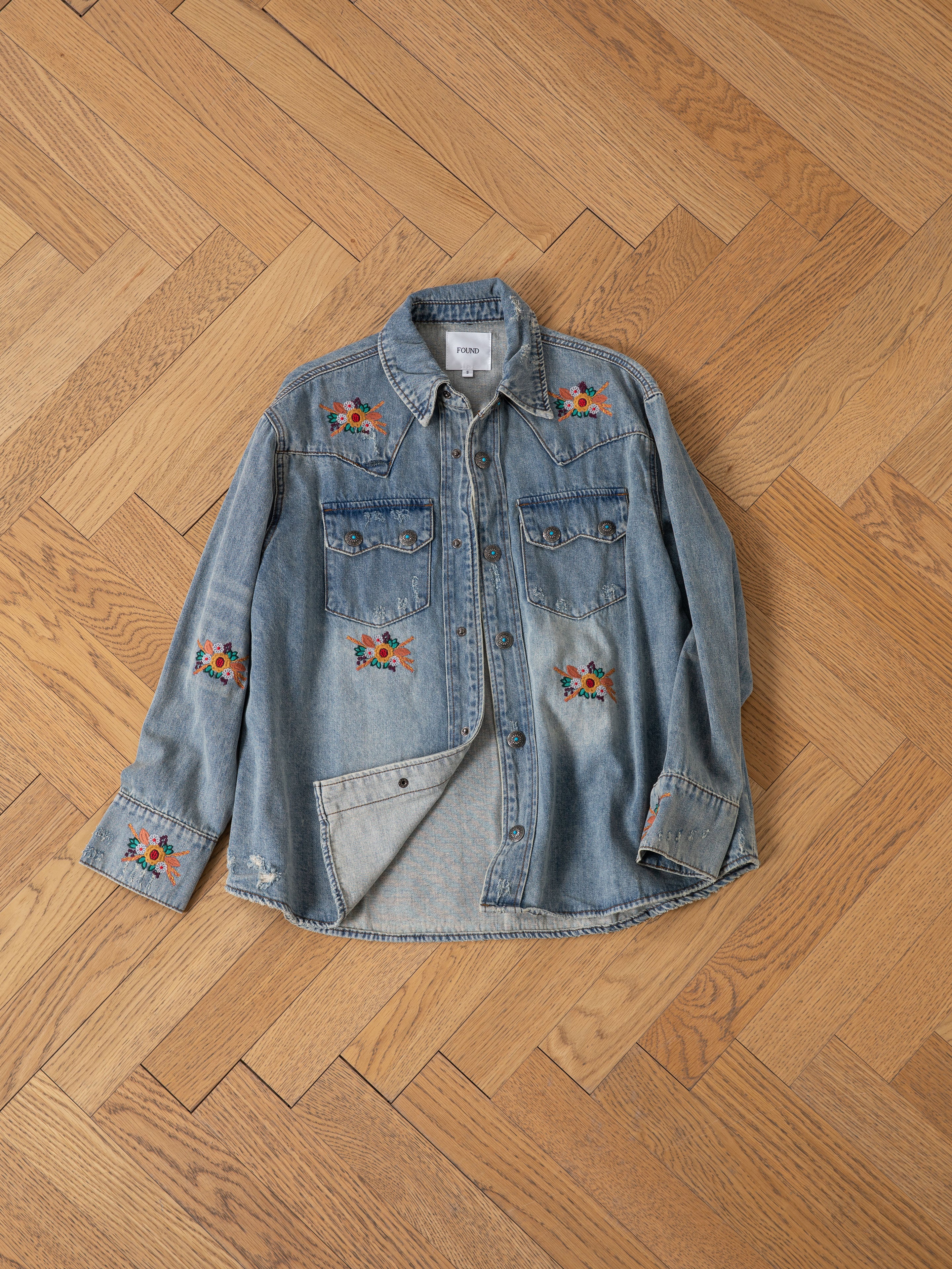 The FOUND Bouquet Floral Embroidered Western Denim Shirt in light blue has a relaxed fit and features floral embroidery on the sleeves, chest, and above the pockets. Displayed on a wooden herringbone floor.