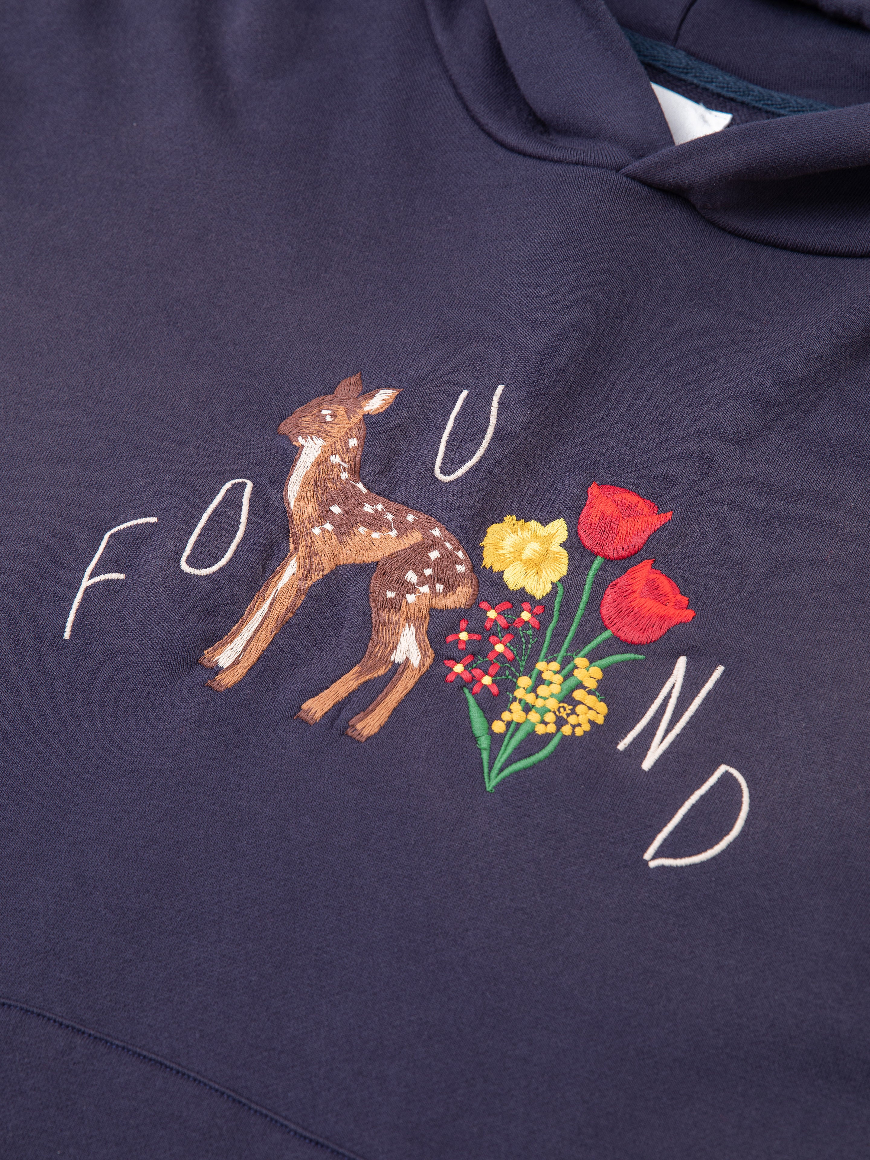 The FOUND Floral Embroidered Deer Hoodie is made from vintage-washed French terry cotton and features intricate embroidery of a fawn with nature-themed elements like yellow, red, and green flowers.