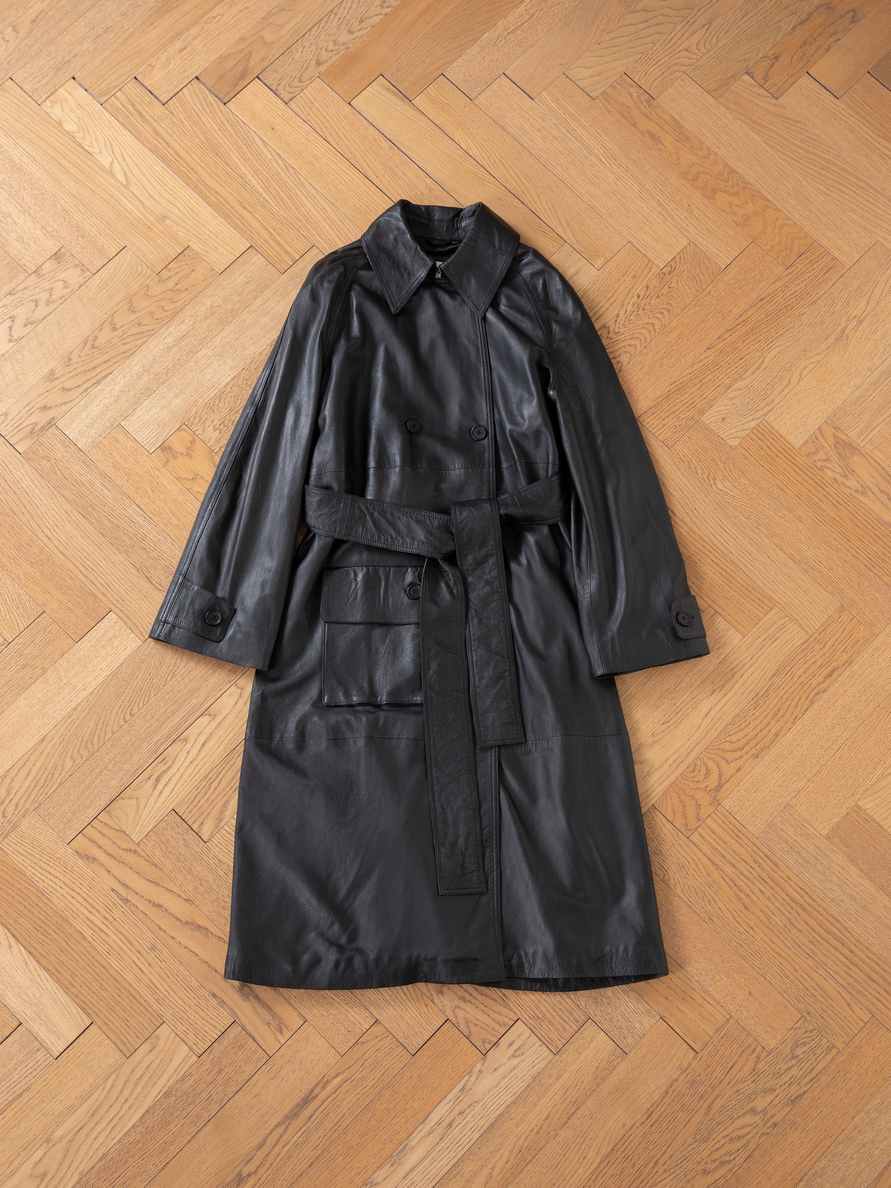The FOUND Leather Trench Coat, an ethically crafted unisex piece with a belt and large pockets, is displayed flat on a light herringbone wooden floor.