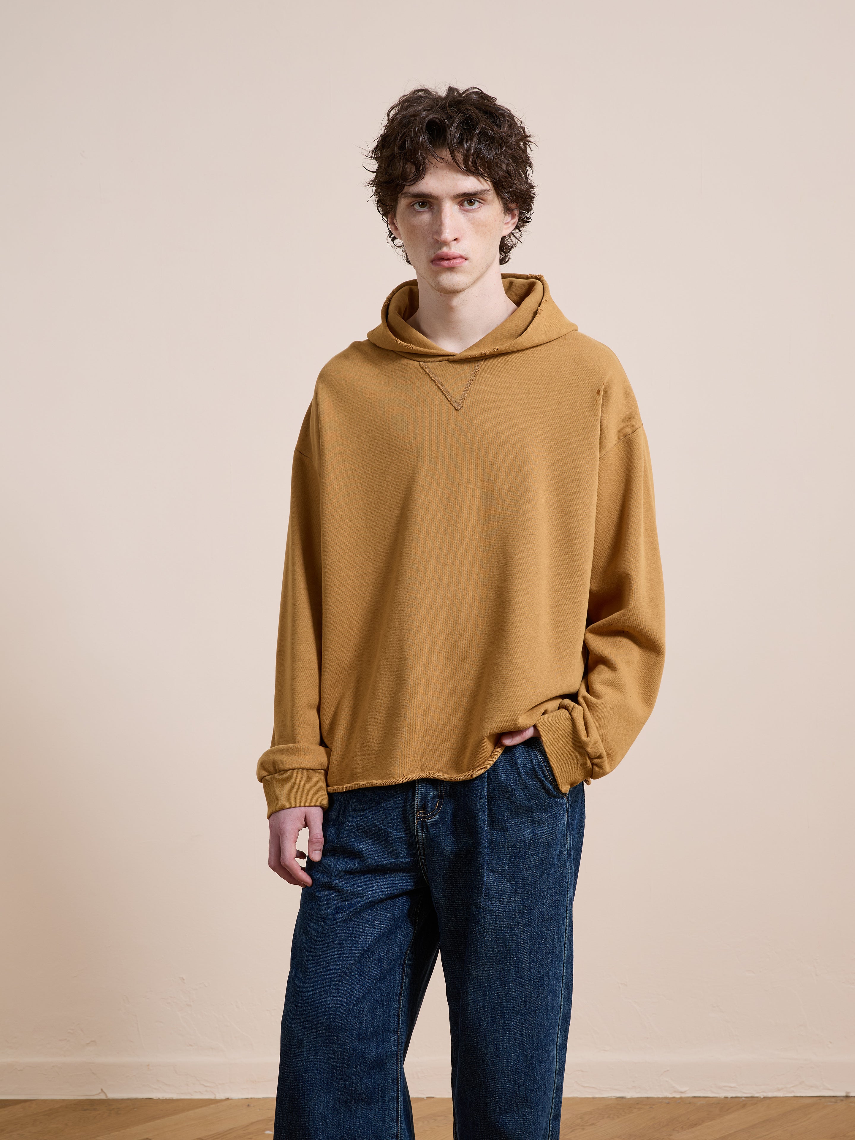 A person wearing the FOUND Beadwork Logo Hoodie in vintage enzyme wash mustard yellow and blue jeans stands against a plain beige wall, looking at the camera.