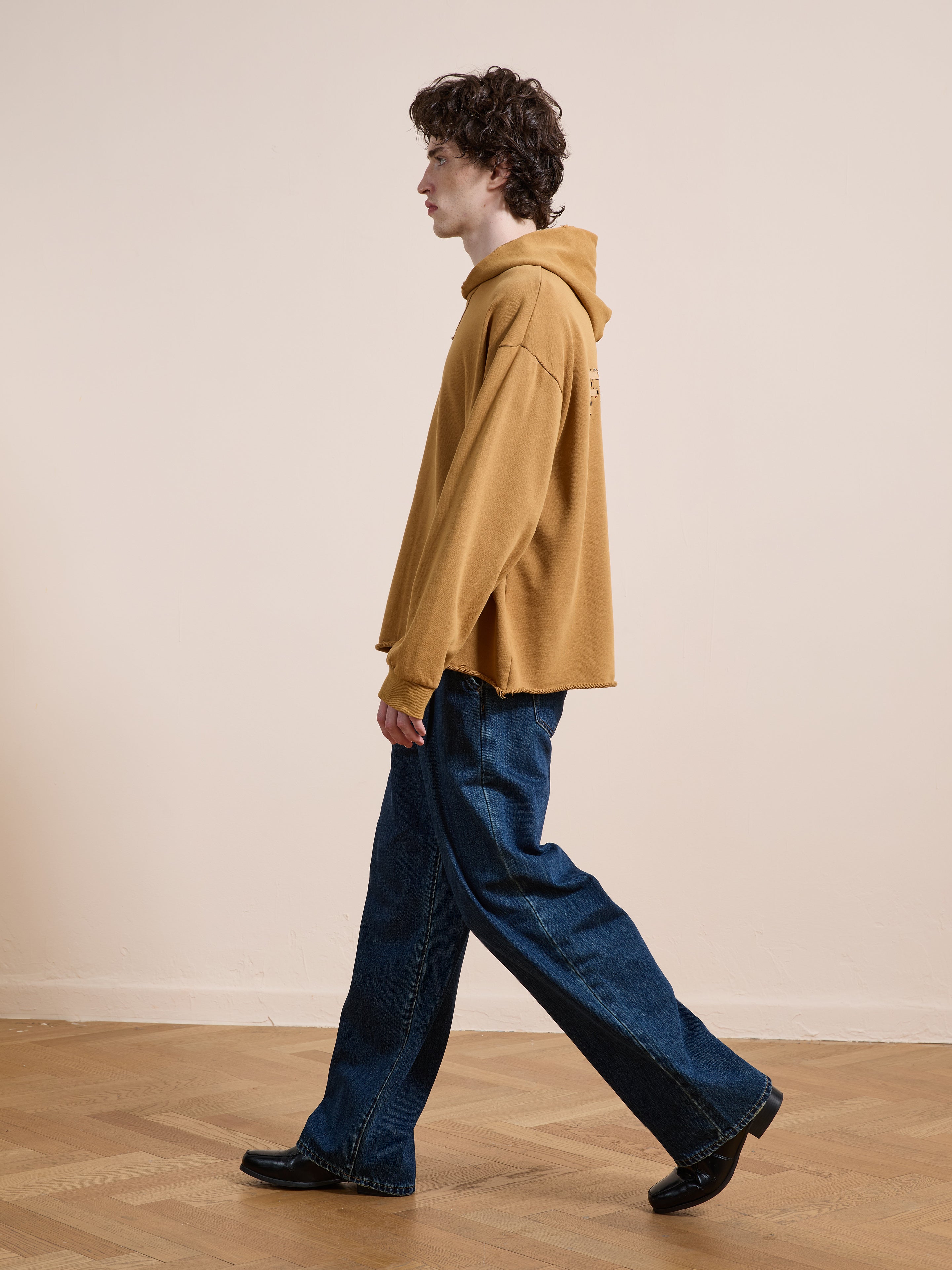 A person in the FOUND Beadwork Logo Hoodie and wide-leg blue jeans walks indoors on a wooden floor, seen in profile against a plain beige wall.