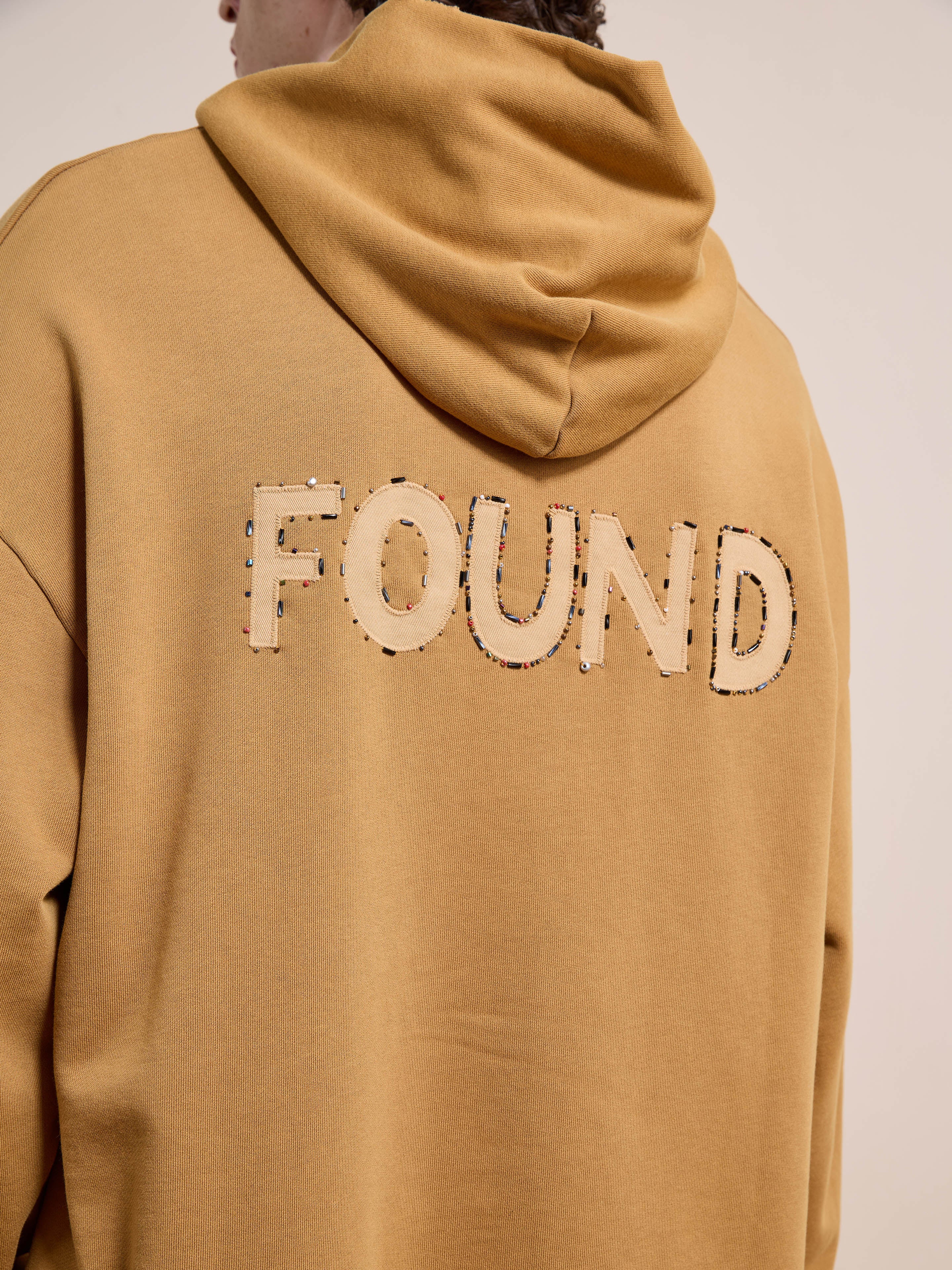 Model wears the FOUND Beadwork Logo Hoodie, a unisex relaxed-fit style in tan french terry with "FOUND" stitched large on the back, finished with a vintage enzyme wash.