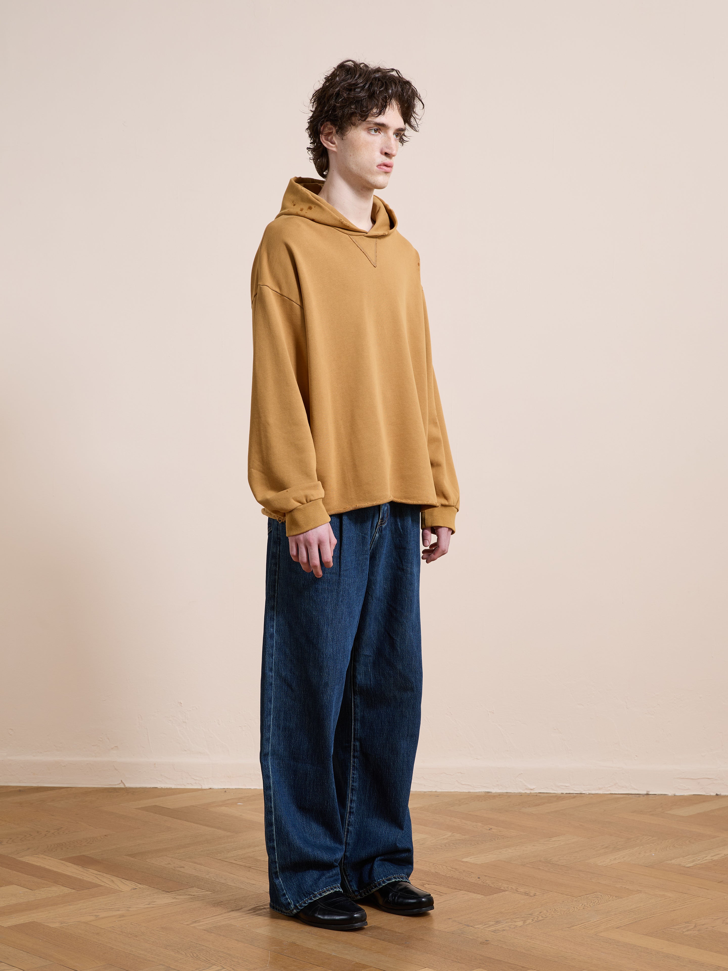 A person stands indoors in a FOUND Beadwork Logo Hoodie, wide-leg blue jeans, and black shoes against a plain beige wall and wooden floor.
