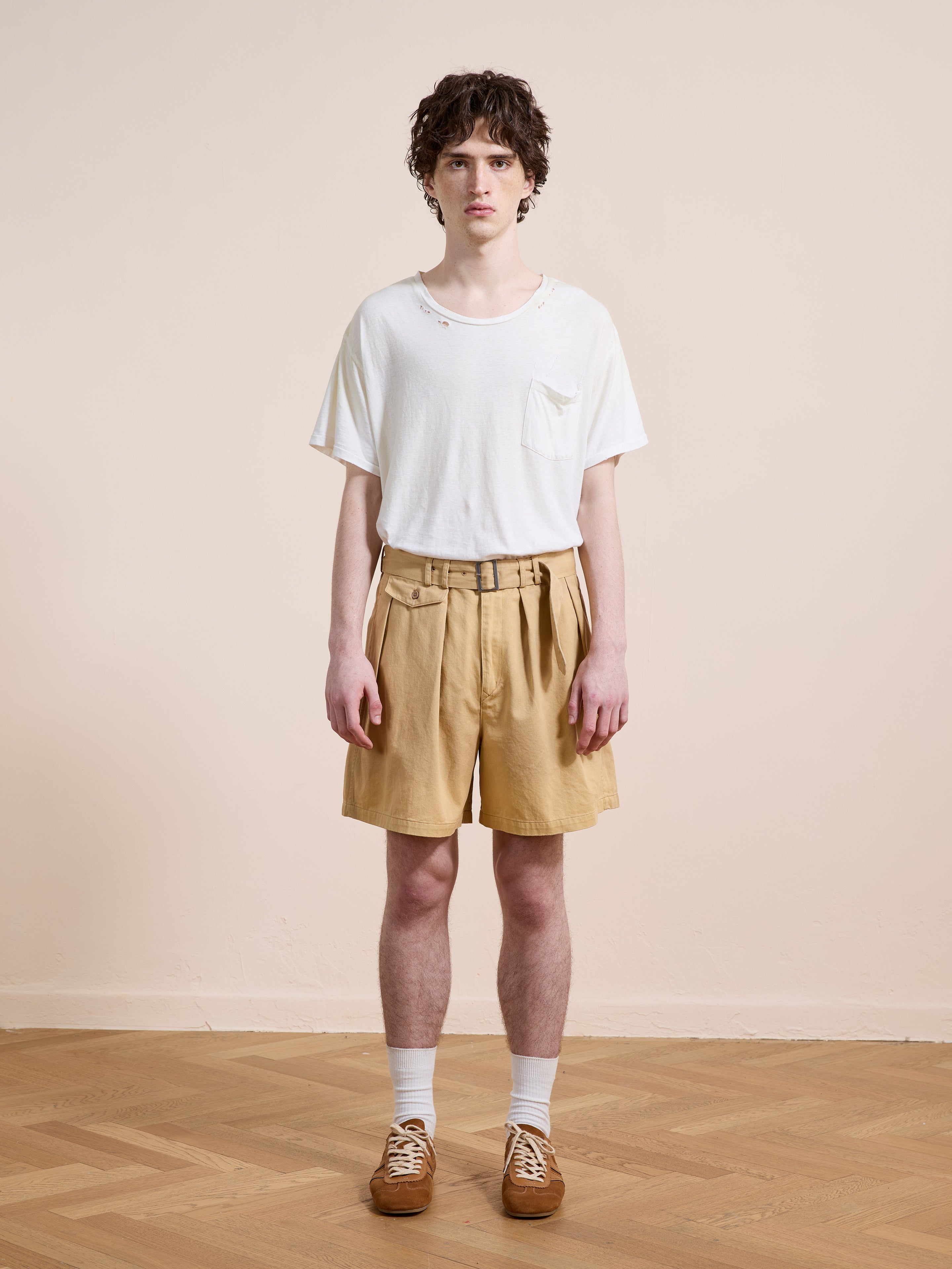 A person stands indoors against a plain wall, wearing a white t-shirt, Found Belted Twill Pleated Shorts in tan, white socks, and brown shoes.