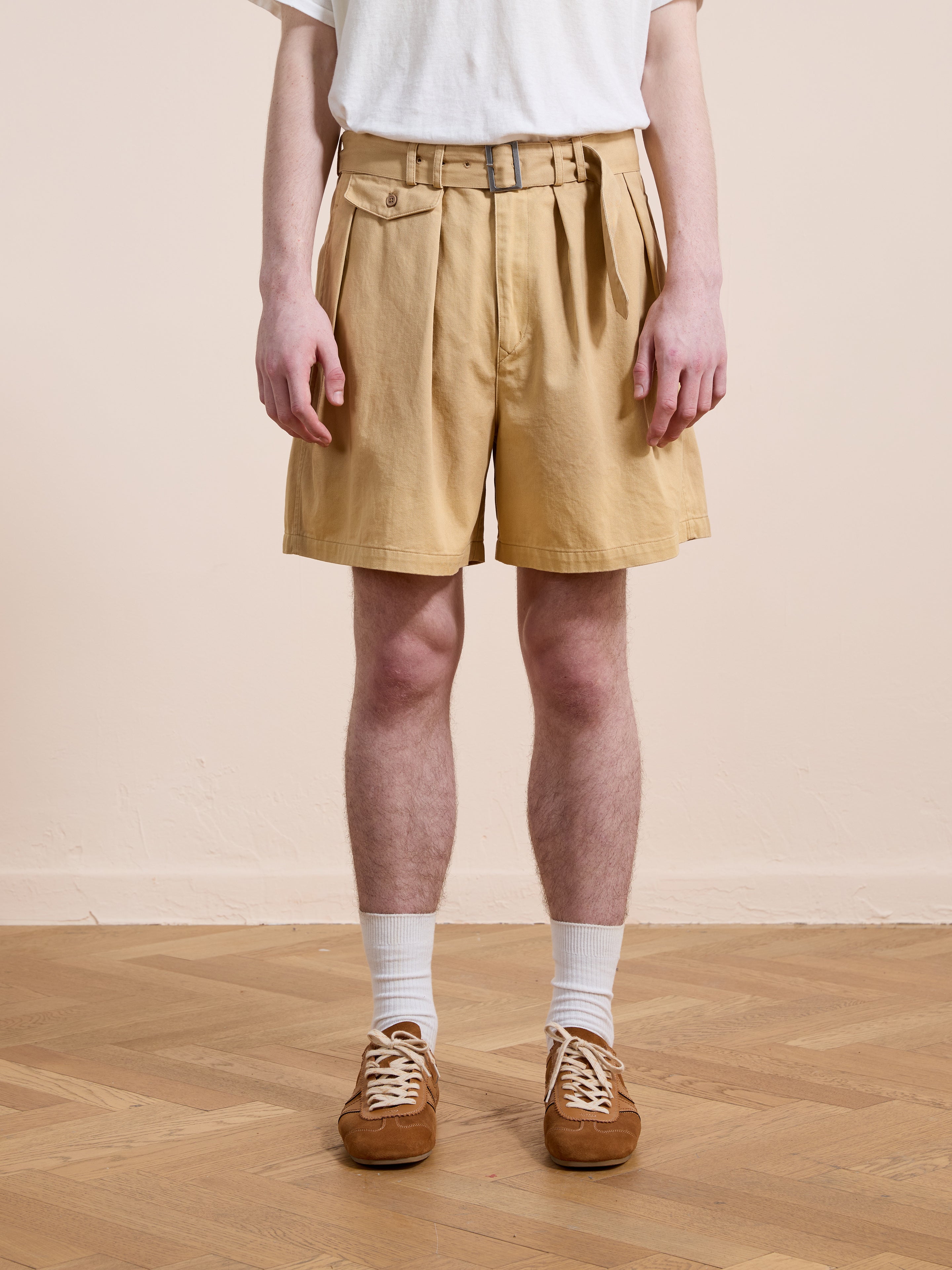 A person stands indoors on a wooden floor wearing a white t-shirt, white socks, brown sneakers, and Found's Belted Twill Pleated Shorts.