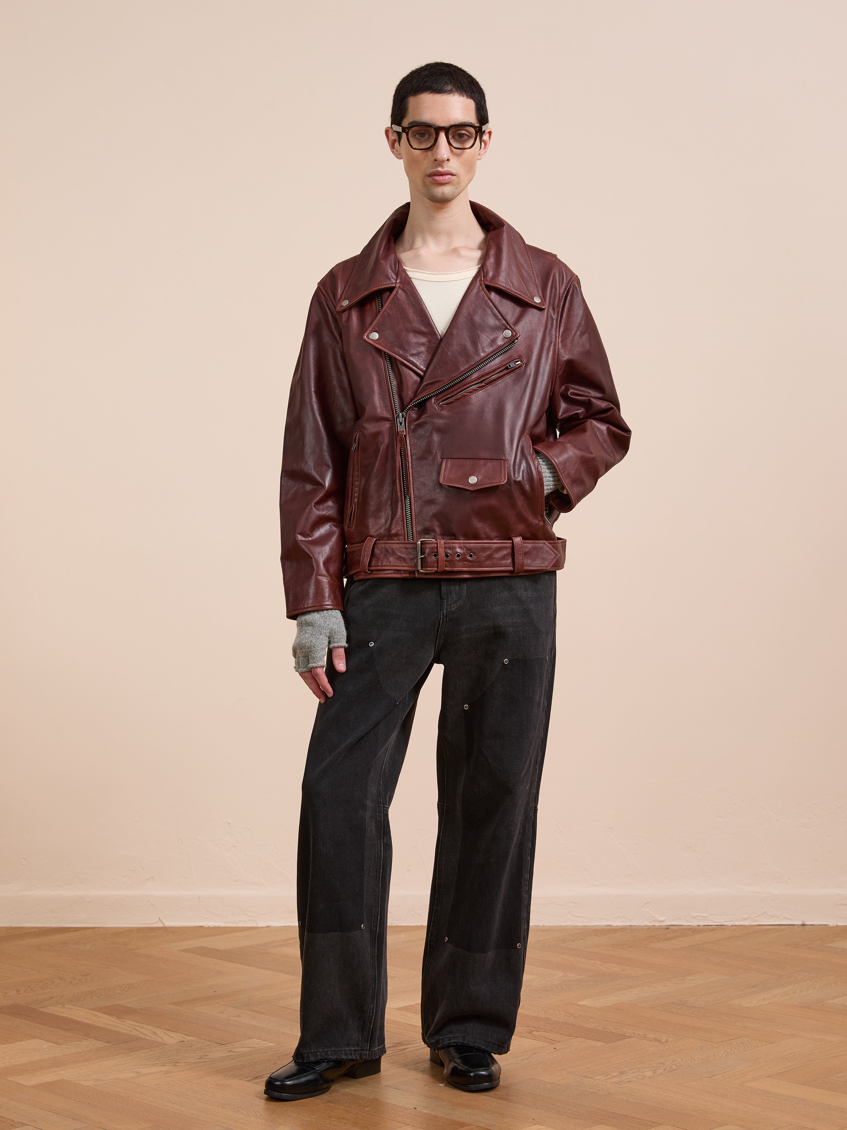 Indoors against a beige background, a person wears FOUND's maroon Leather Biker Jacket, paired with a white shirt, black wide-leg pants, black shoes, gray gloves, and glasses.