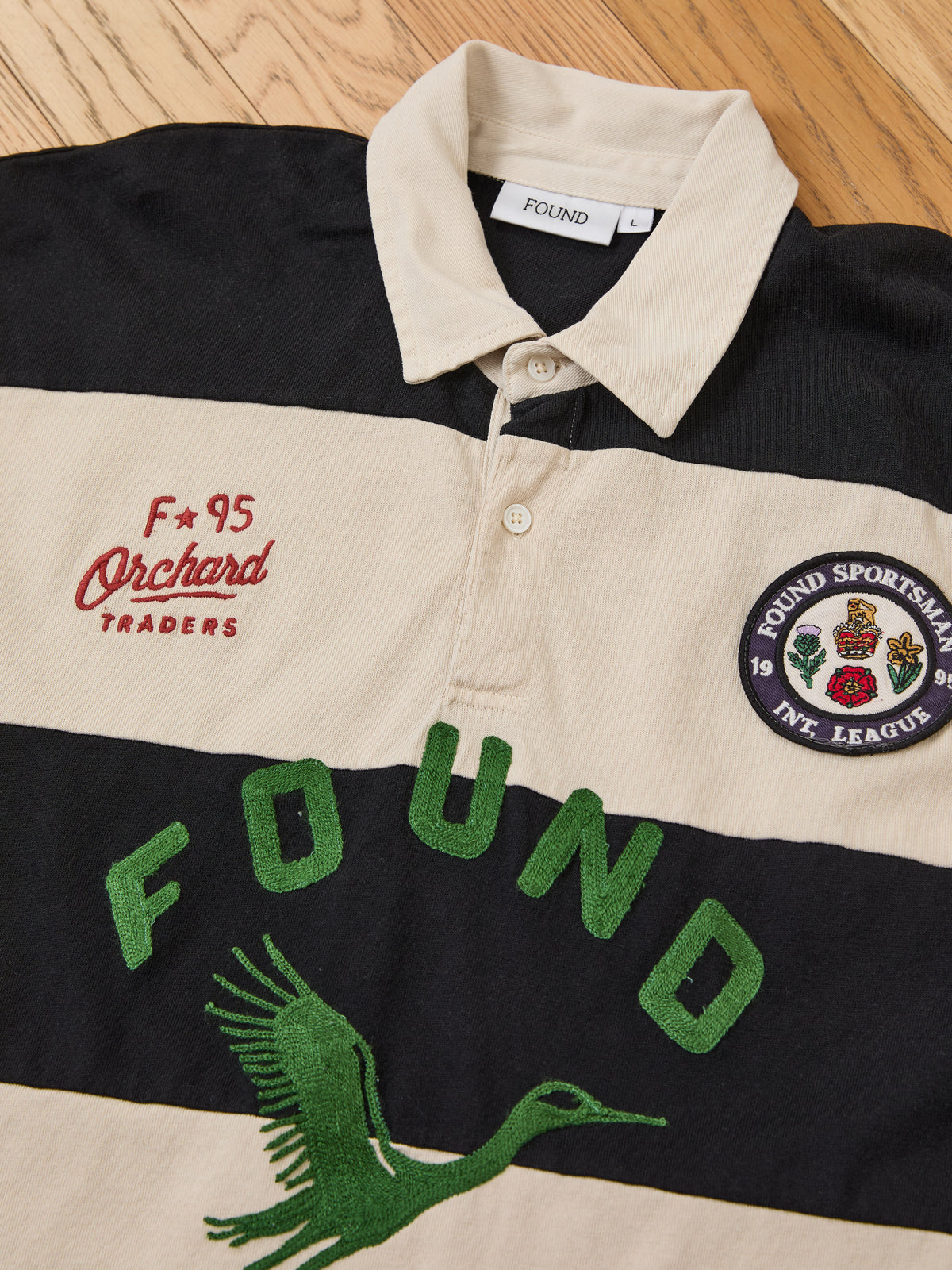 The Bird Logo Rugby by FOUND is a black and cream cotton top with “FOUND,” a bird, “Orchard Traders,” and crest patches in a unisex relaxed fit.