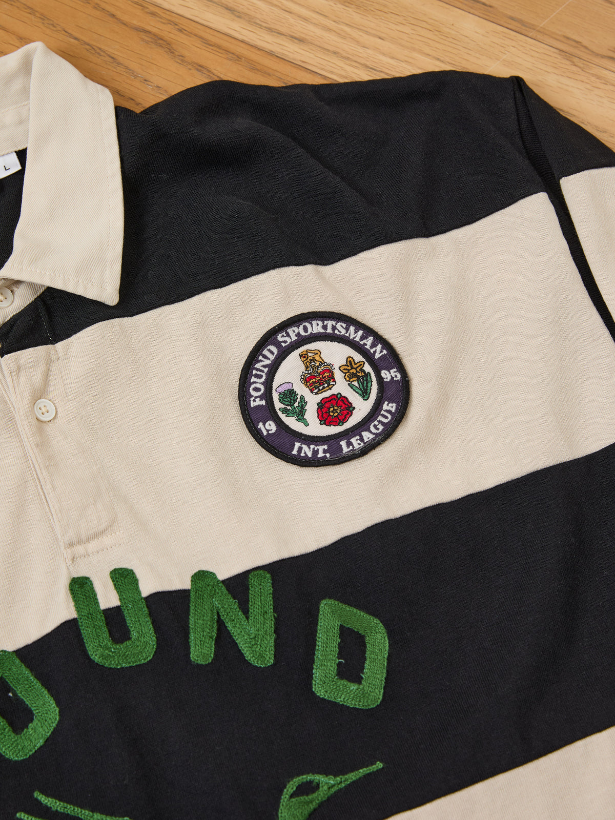 The FOUND Bird Logo Rugby is a black and beige striped cotton top with a patch and green embroidered letters, unisex relaxed fit, shown on a wooden floor.
