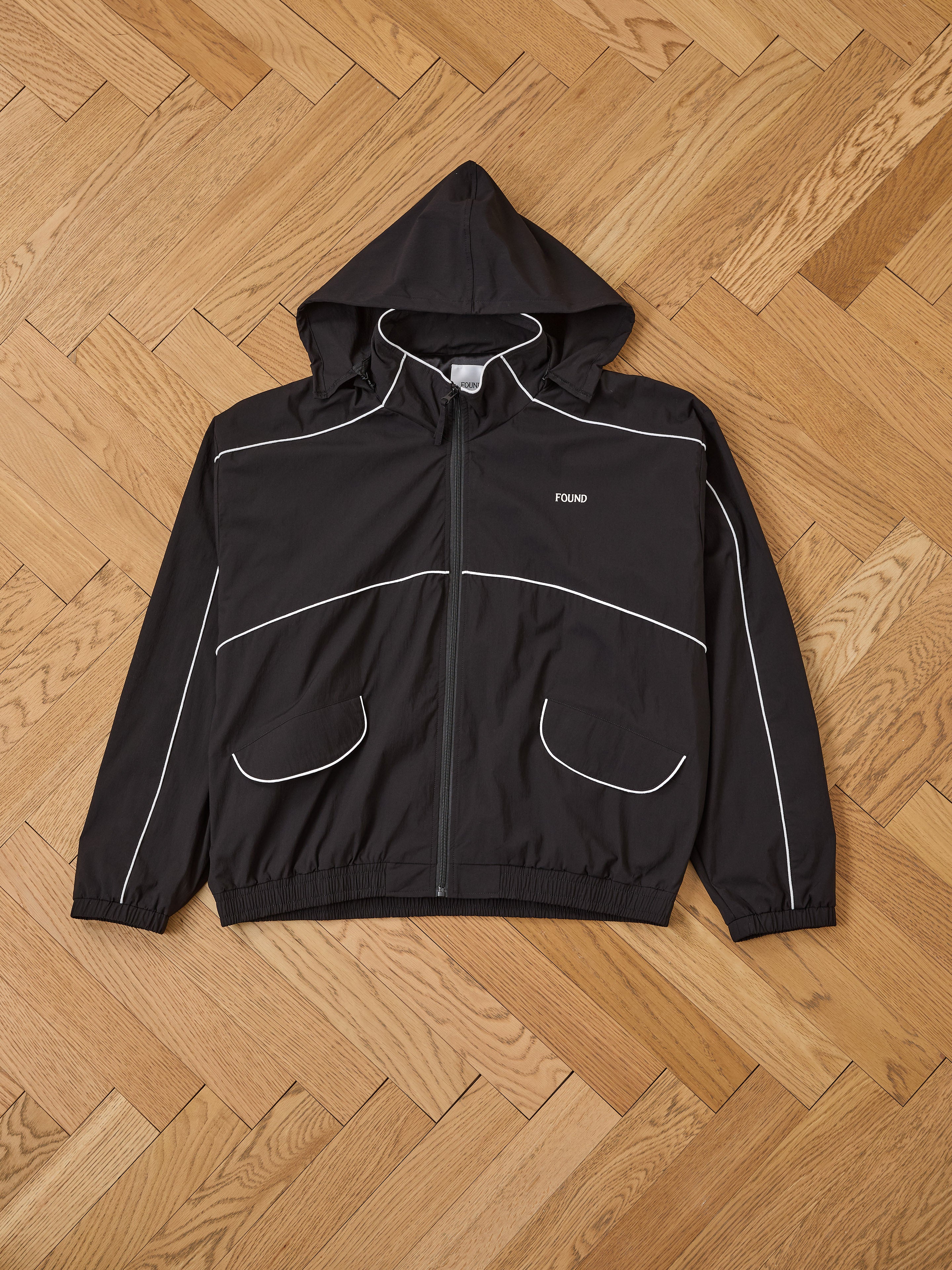 The FOUND Arena Stripe Track Jacket is a black unisex hooded jacket in lightweight technical fabric, with white piping, two front pockets, and "FOUND" logo on the chest, shown laid flat on a light wood herringbone floor.