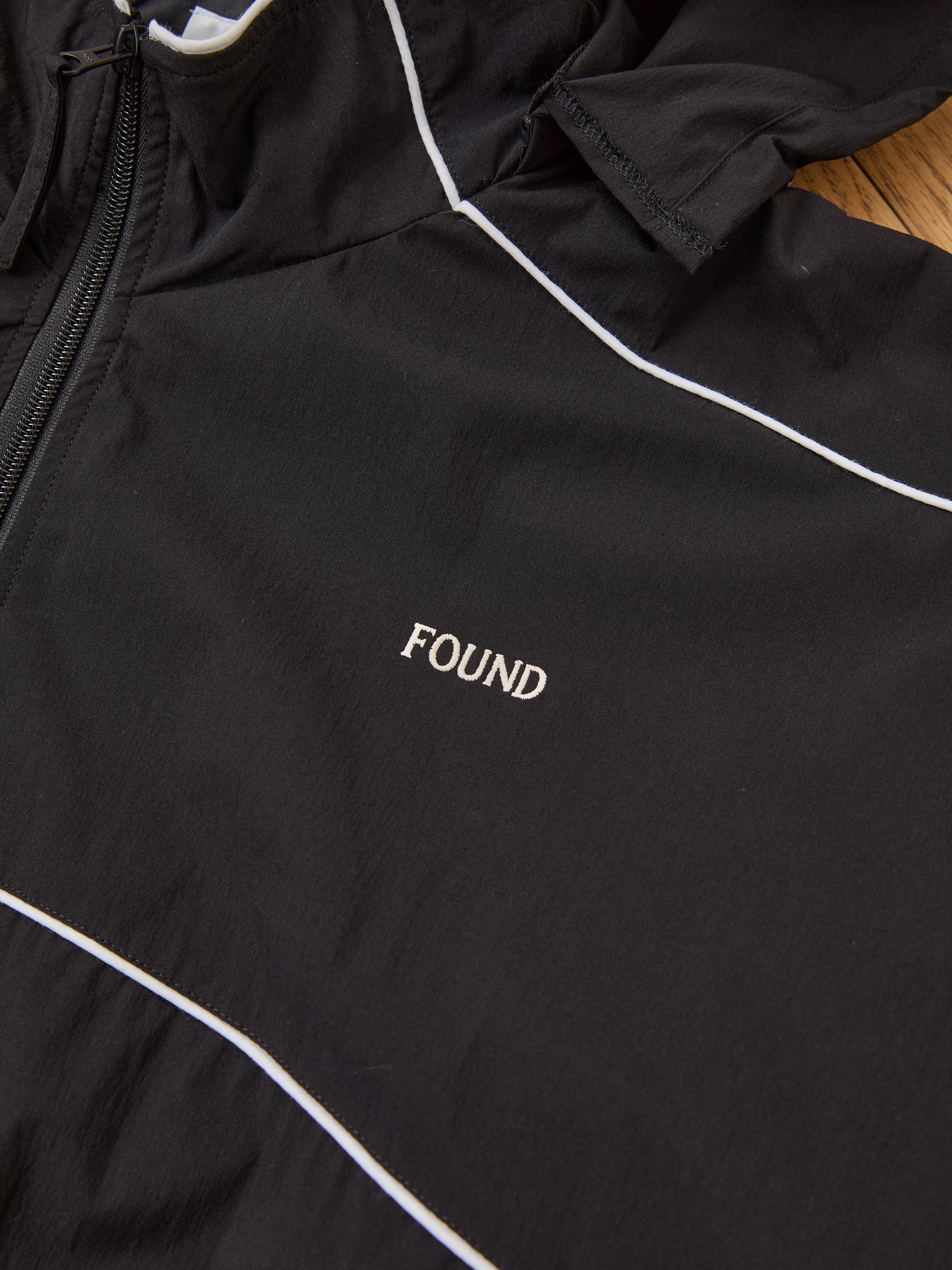 The FOUND Arena Stripe Track Jacket is a unisex black jacket in lightweight technical fabric, featuring white piping and "FOUND" embroidered in white on the chest.