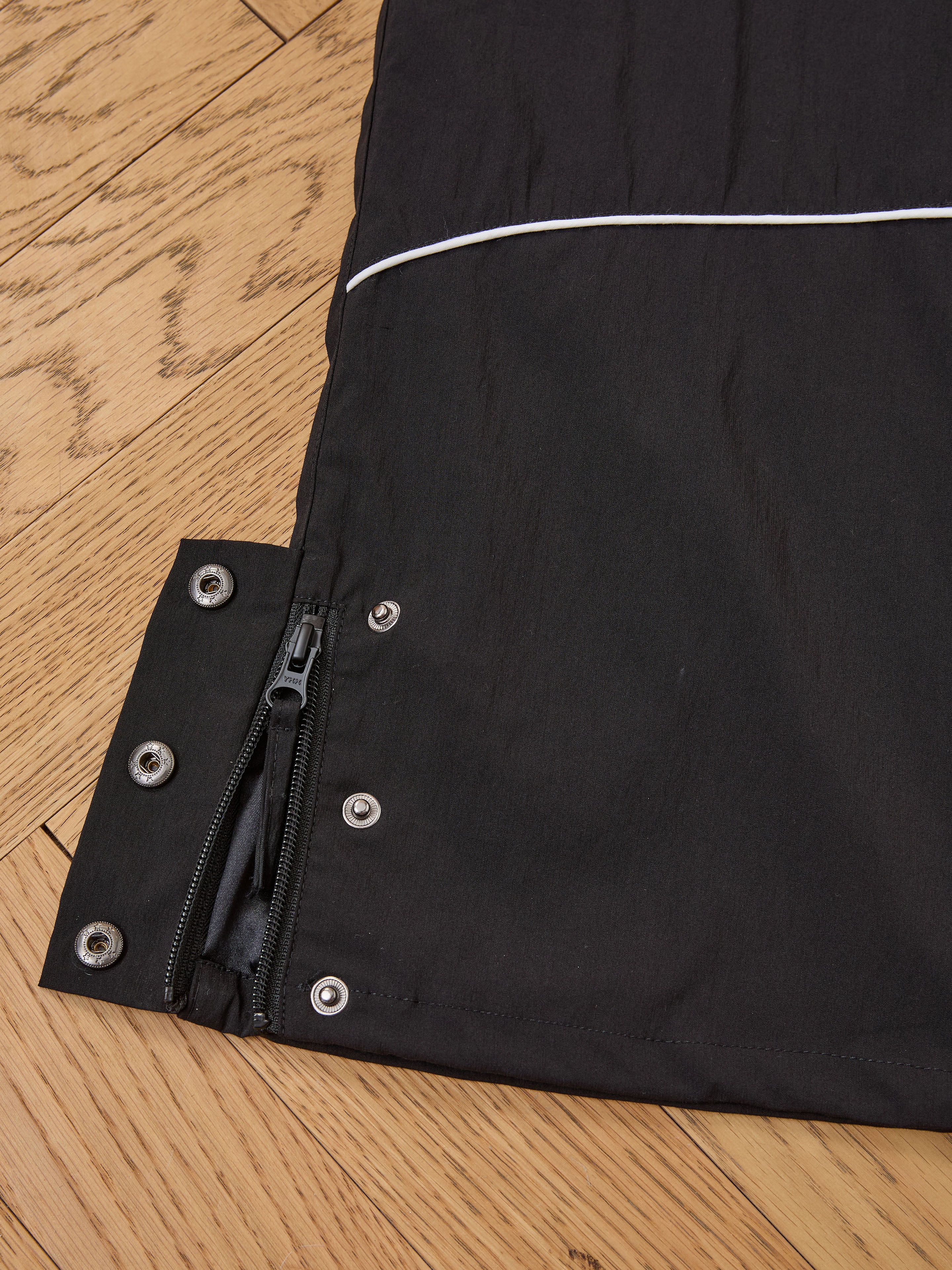 Close-up of Found's Arena Stripe Track Pants sleeve in black, featuring a zipper, snap buttons, and white piping detail on a wooden floor. Classic unisex sportswear with an adjustable waistband for comfort.