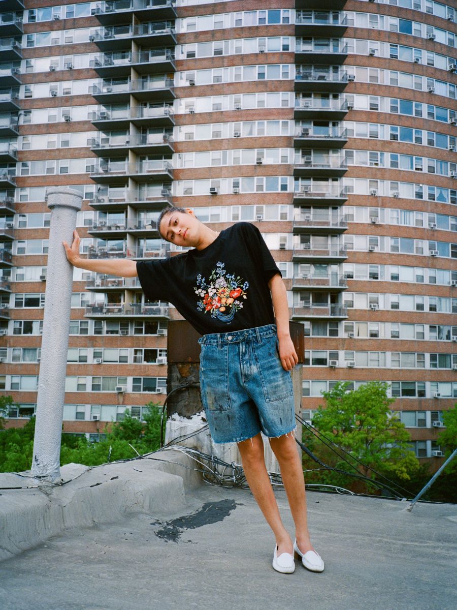 Wearing the FOUND Bouquet Flowers Tee and denim shorts, a person stands on a rooftop, leaning against a white pipe with a tall apartment building in the background.