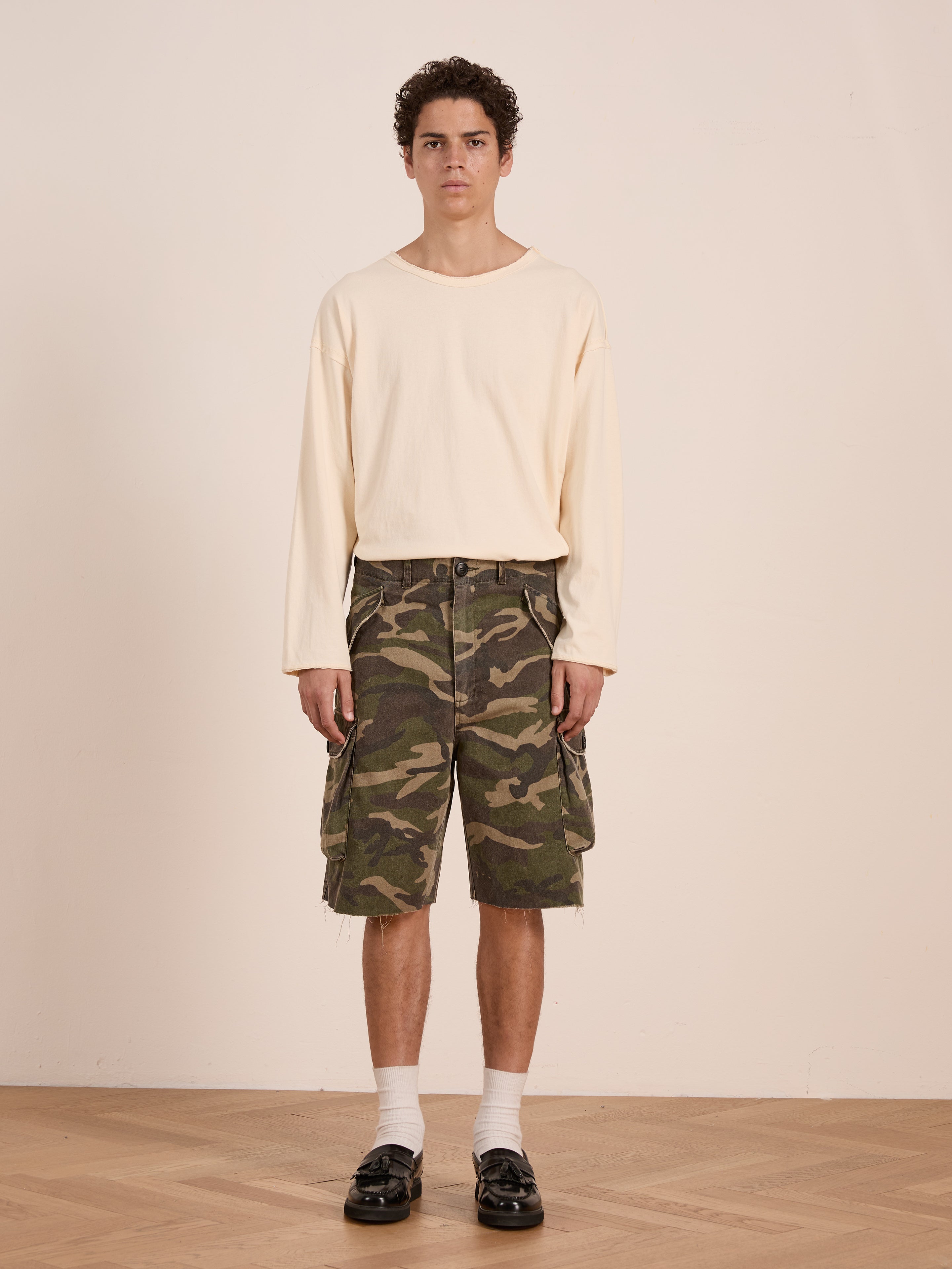 A young man faces the camera indoors, wearing FOUND Camouflage Twill Utility Cargo Shorts and a cream long-sleeve shirt, styled with white socks and black loafers against a plain background for a vintage look.