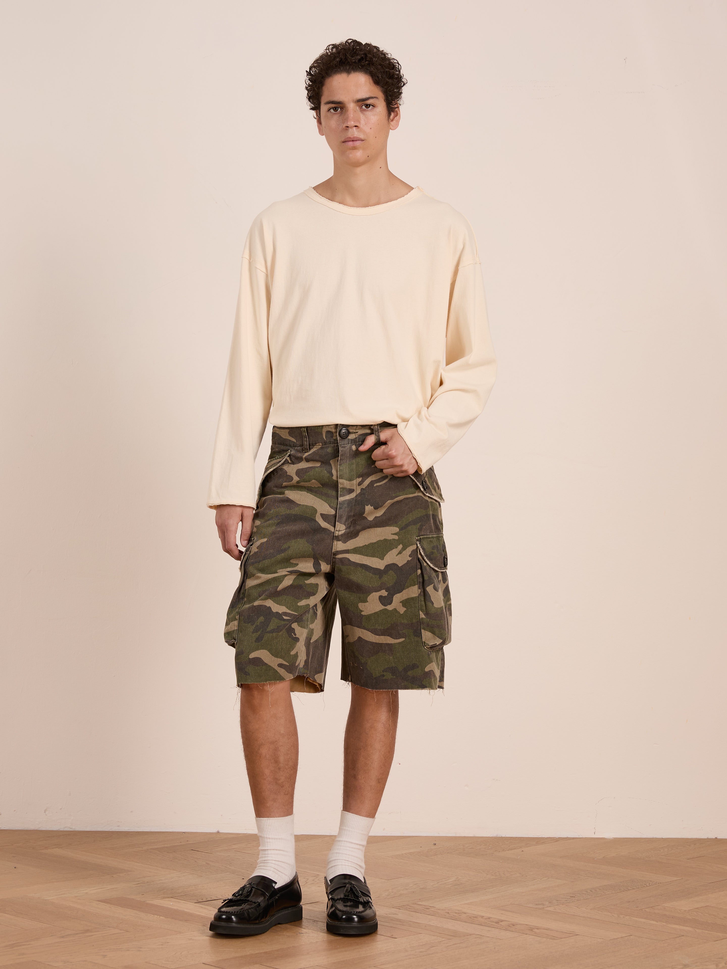 A person stands indoors against a plain background, wearing FOUND Camouflage Twill Utility Cargo Shorts, a loose cream long-sleeve shirt, white socks, and black loafers.