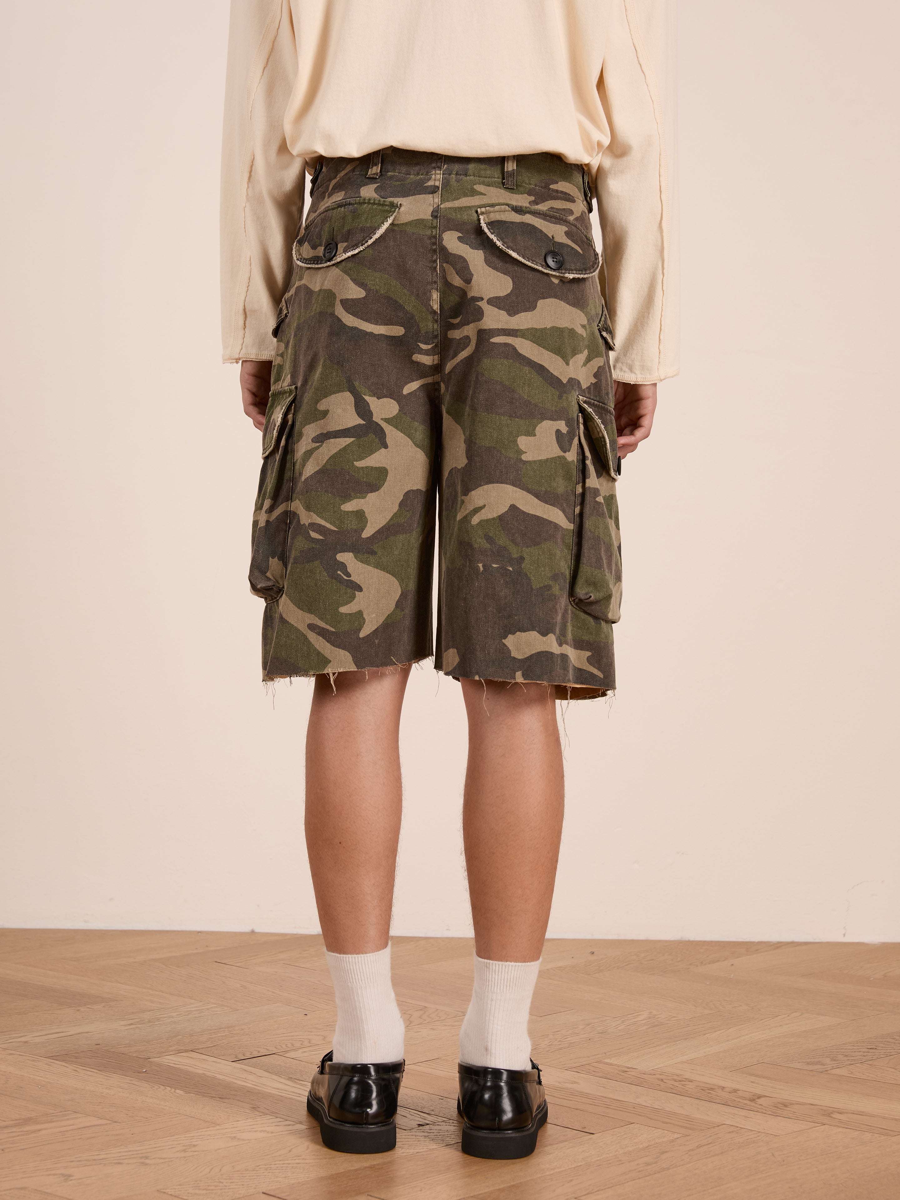 Person wearing FOUND Camouflage Twill Utility Cargo Shorts, featuring a vintage look and unisex relaxed fit for style and comfort, paired with white socks and black shoes on a wooden floor, shown from the back.