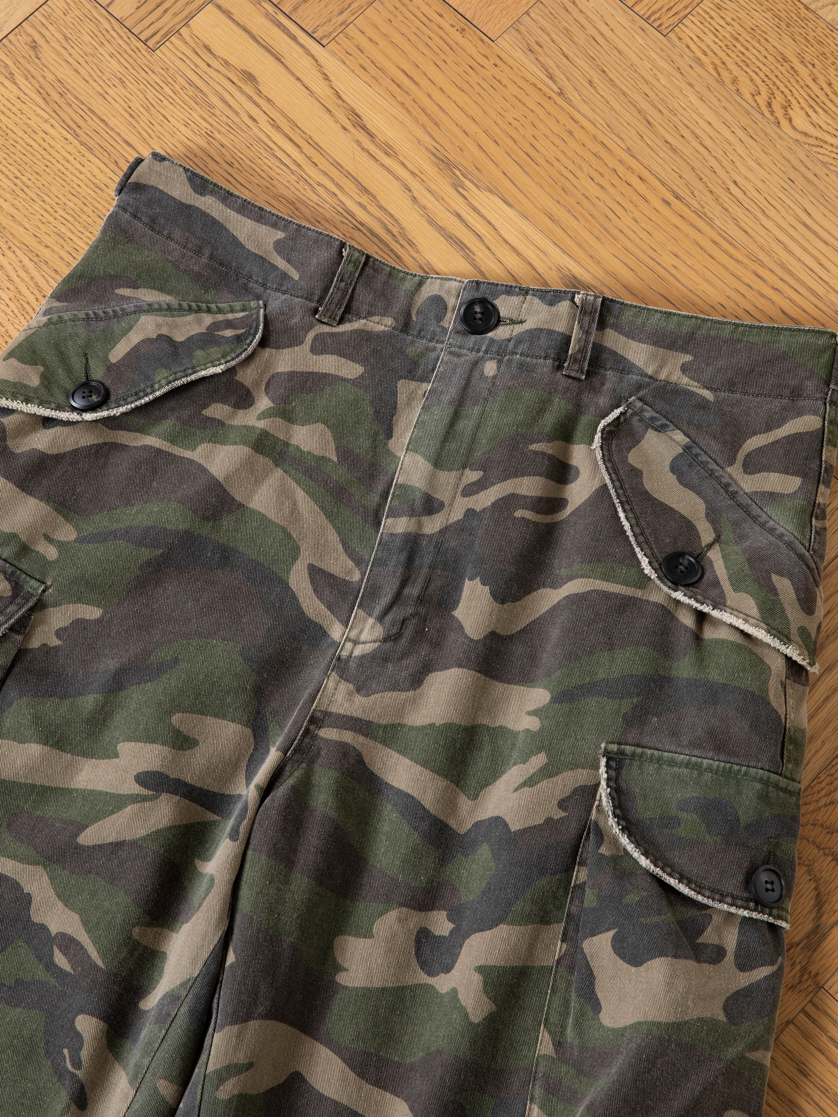 A pair of FOUND Camouflage Twill Utility Cargo Shorts with buttoned flap pockets, featuring a vintage look, is displayed on a wooden parquet floor.