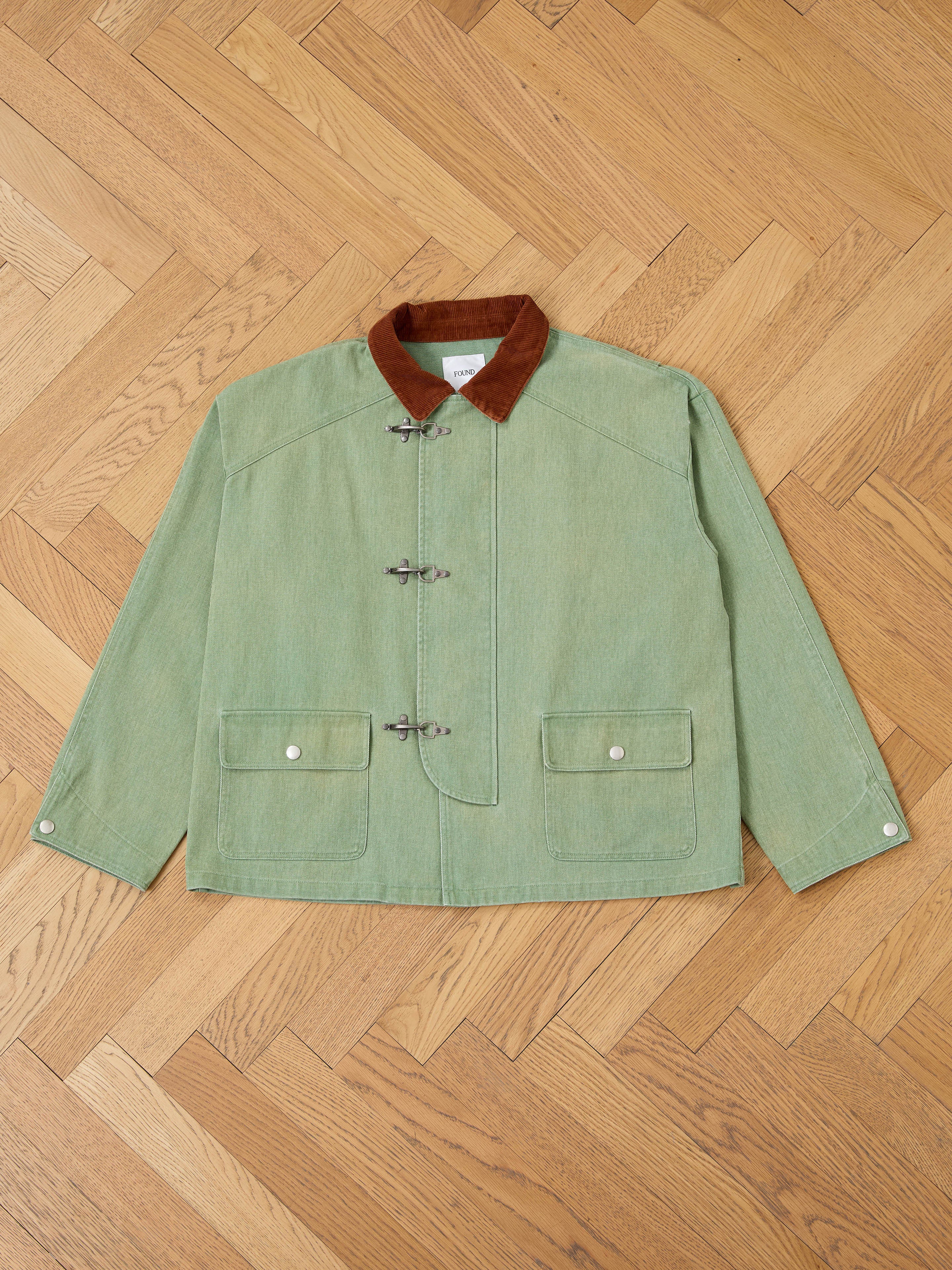 The Found Washed Fireman Clasp Workshop Jacket, a green unisex piece with a corduroy collar, toggle closures, and two front flap pockets, is laid flat on a wooden parquet floor.