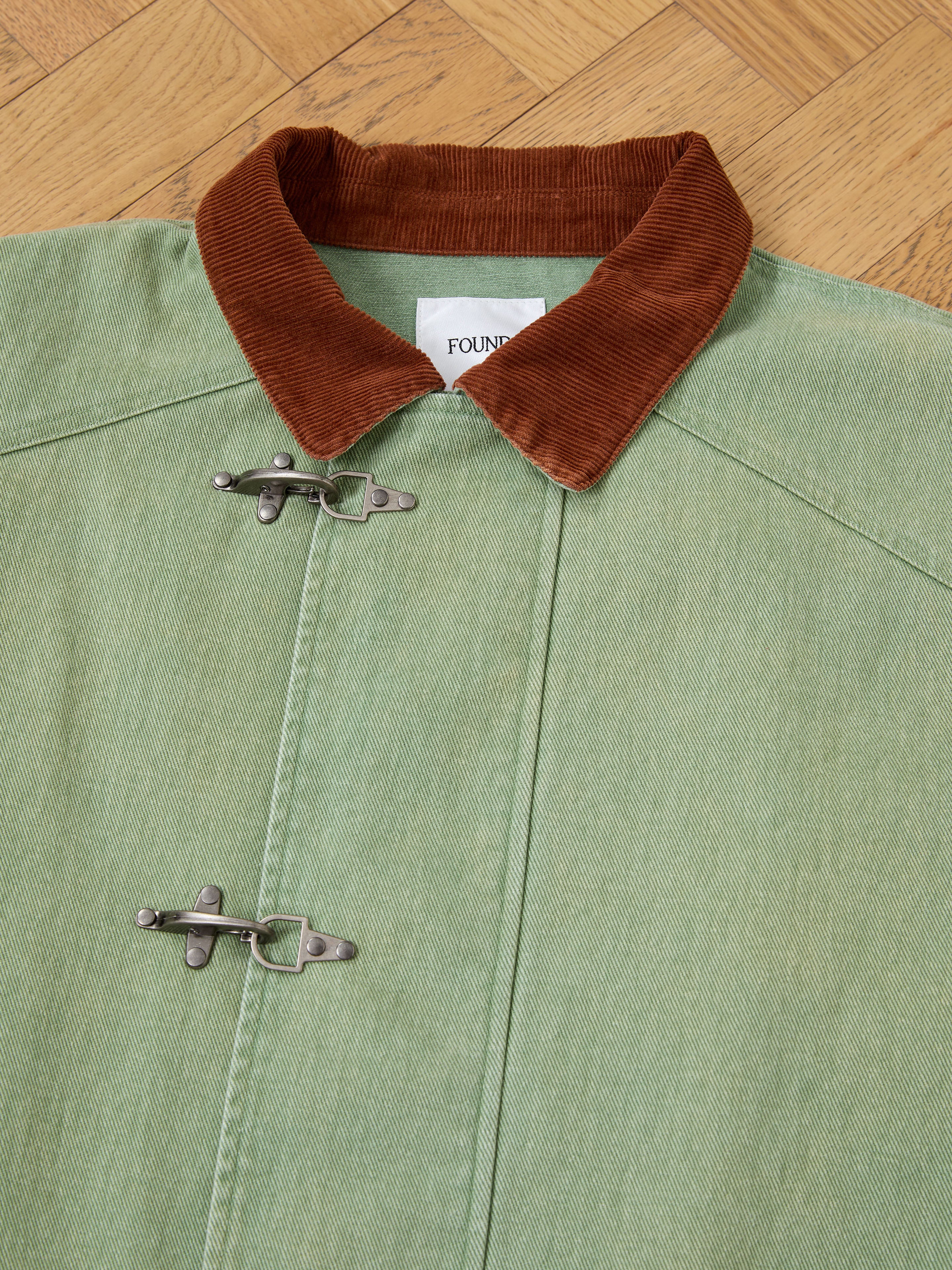 A close-up of the Found Washed Fireman Clasp Workshop Jacket, a unisex utility piece with a brown corduroy collar and metal clasp fasteners, shown laid flat on a wooden floor.