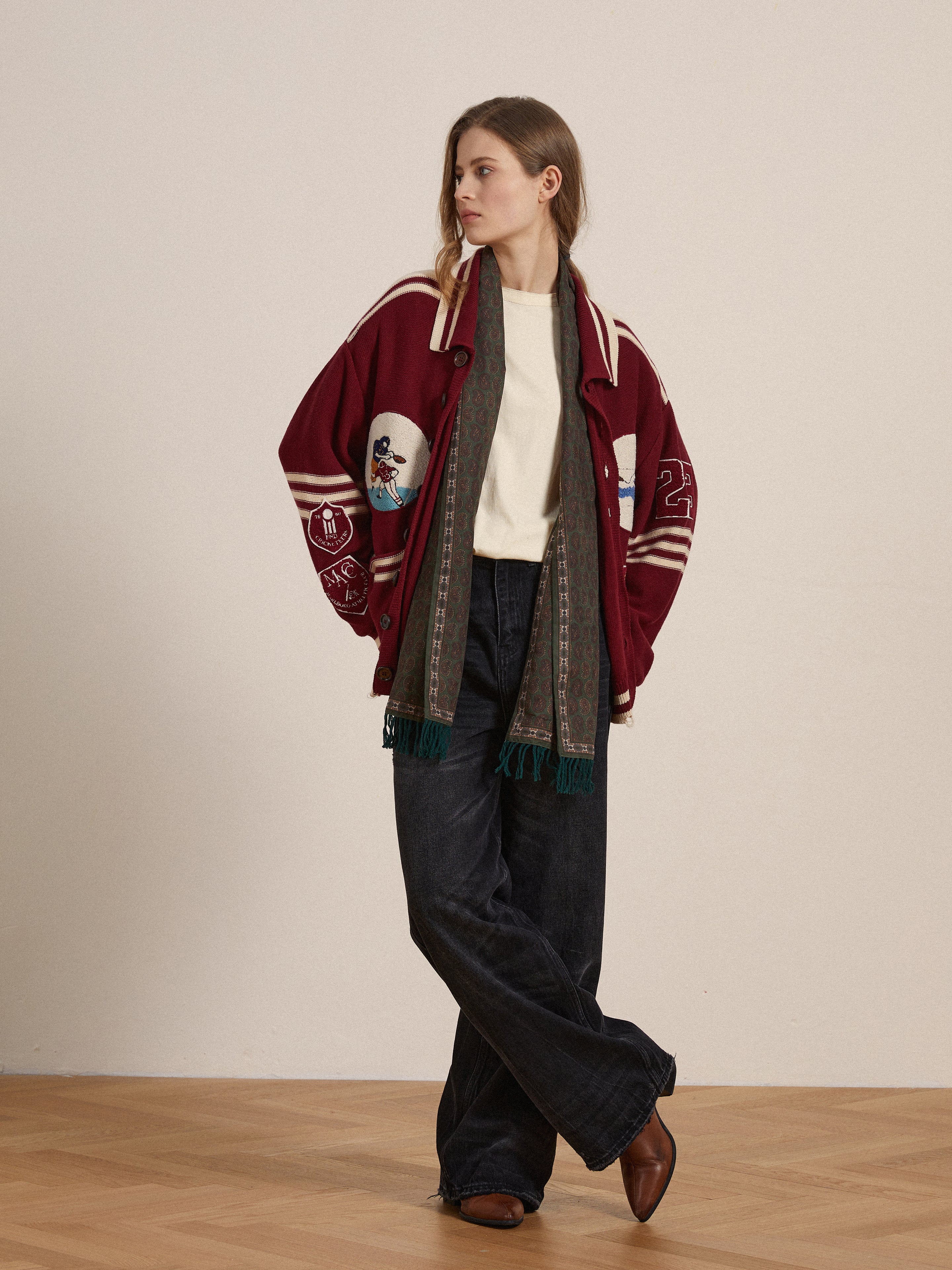 A woman stands indoors on a wooden floor against a plain wall, wearing FOUND's Reversed LS Tee with a distressed neckline under a red varsity jacket, styled with a patterned scarf, wide-leg black jeans, and brown shoes.