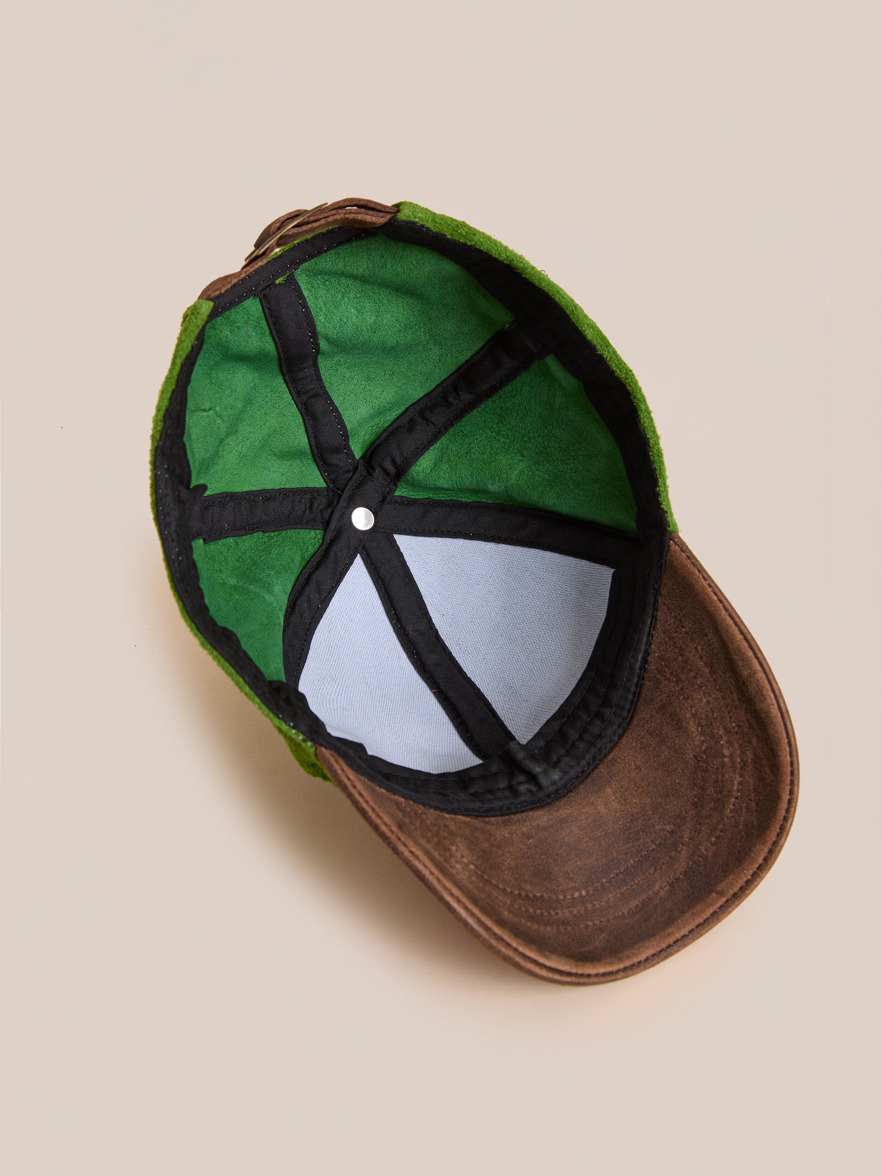 The Suede Leather Contrast Distressed Cap by Found is pictured upside down, revealing its green suede exterior, fabric interior, black stitched panels, and distressed leather brim against a plain beige background.