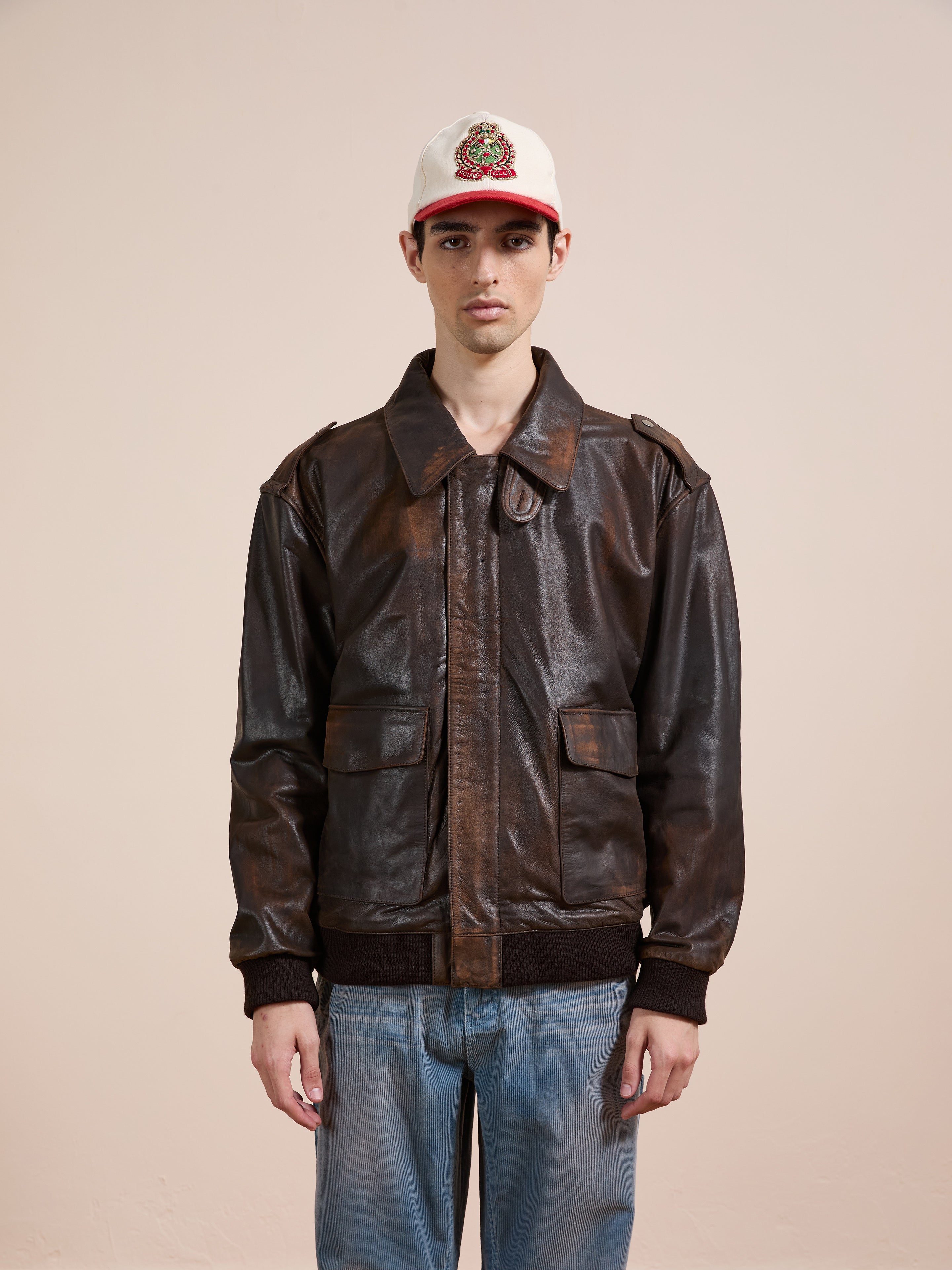 A person wearing the FOUND Distressed Leather Flight Jacket, blue jeans, and a white baseball cap stands against a plain beige background.
