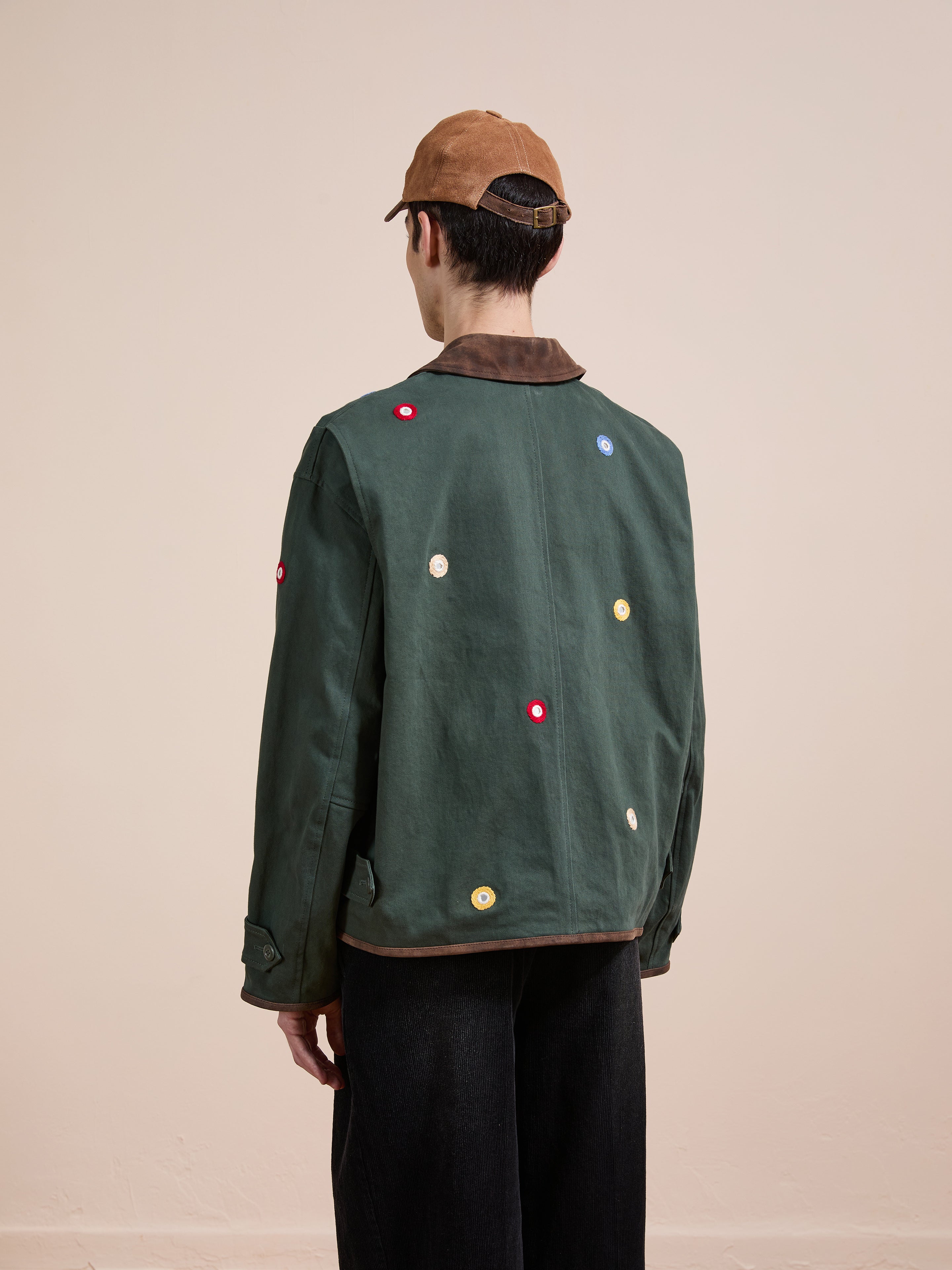 A person stands with their back to the camera against a plain background, wearing black pants, a brown cap, and the Found Hand Embroidered Mirror Abhala Jacket in green with colorful circular patches.