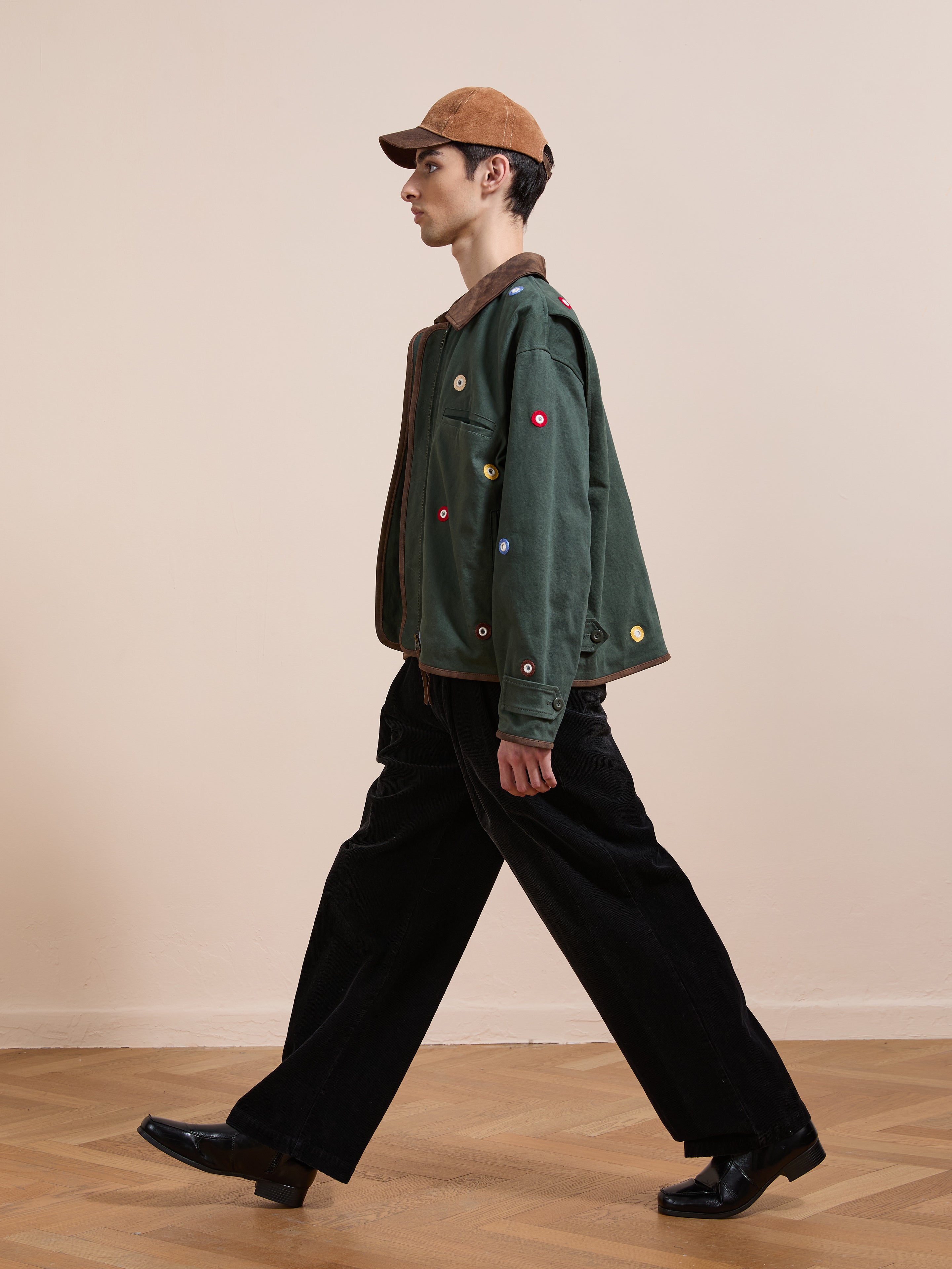 A person in a brown cap, Found’s Hand Embroidered Mirror Abhala Jacket, black wide-leg pants, and black shoes walks on a wooden floor with a plain beige background.