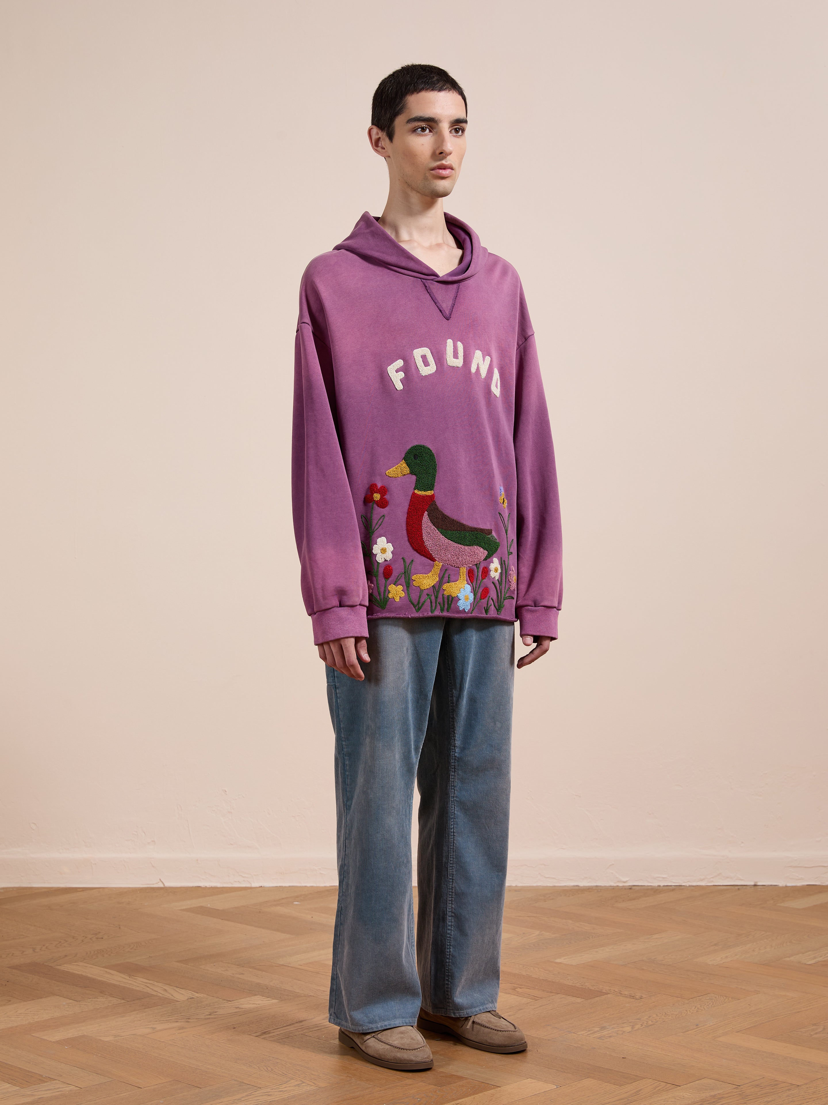 Someone stands indoors on a wooden floor wearing the FOUND Embroidered Wildlife Hoodie in purple, featuring a duck graphic, paired with blue wide-leg jeans and beige shoes.