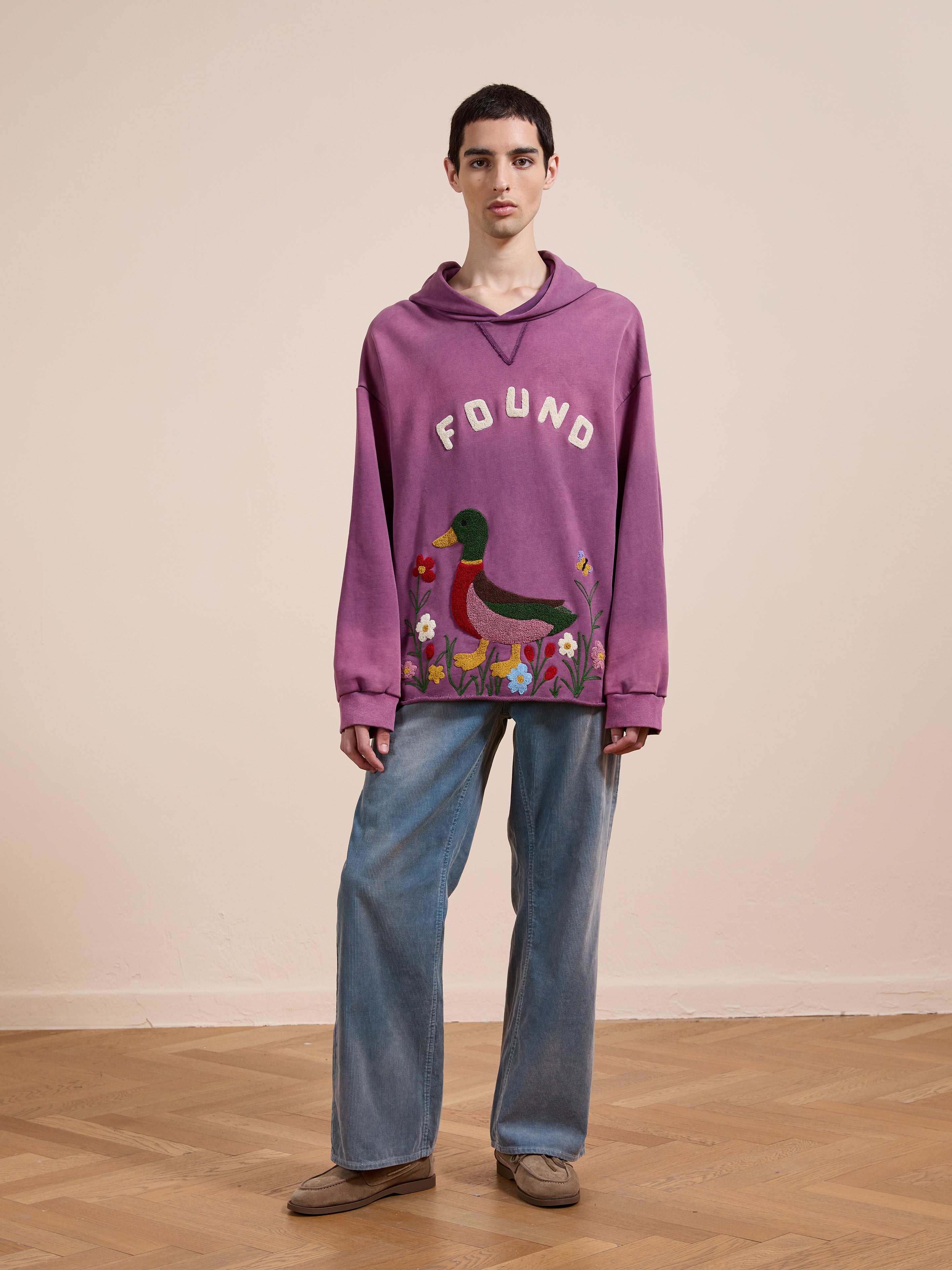 Indoors, a person wears the FOUND Embroidered Wildlife Hoodie in purple, featuring a colorful duck graphic, styled with wide-leg jeans and brown shoes, against a beige wall and wooden floor.