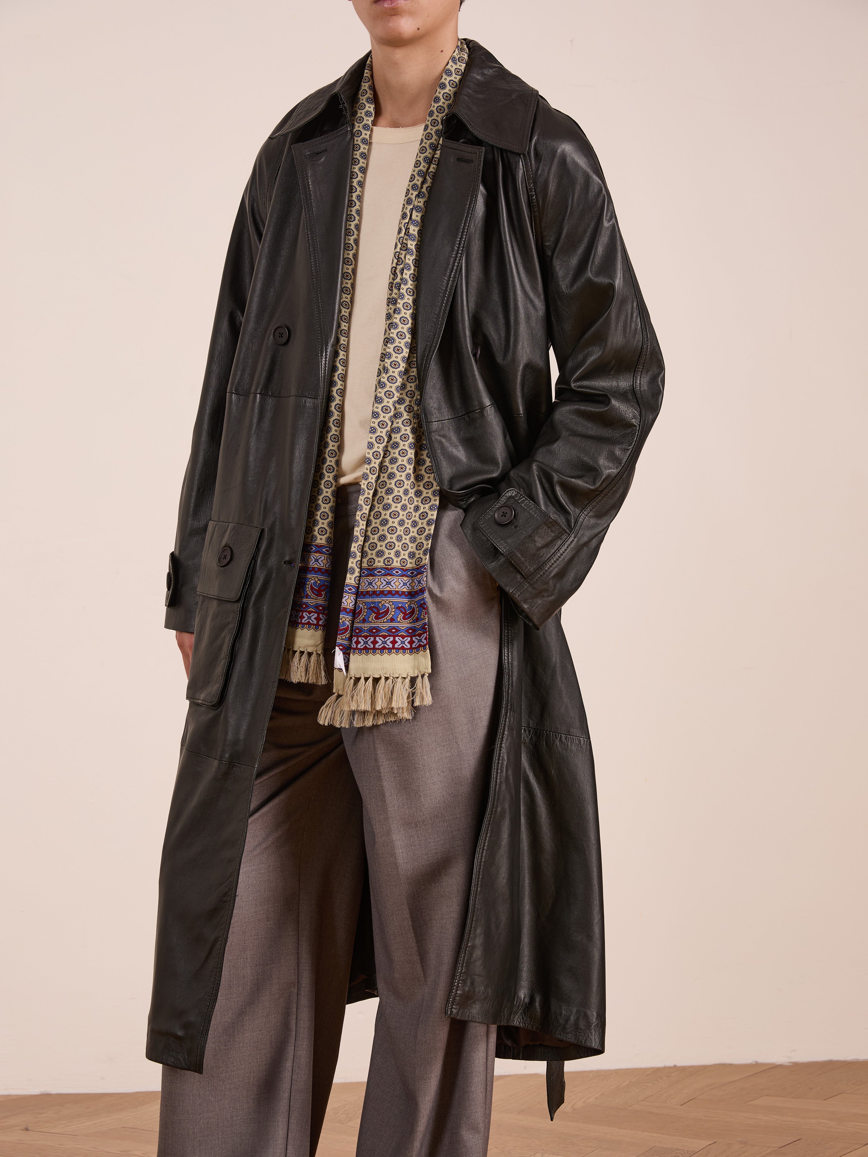 A person wearing the FOUND Leather Trench Coat—ethically crafted from lightweight black cowhide—styled with a light shirt, fringed patterned scarf, and gray wide-leg trousers stands against a plain background.