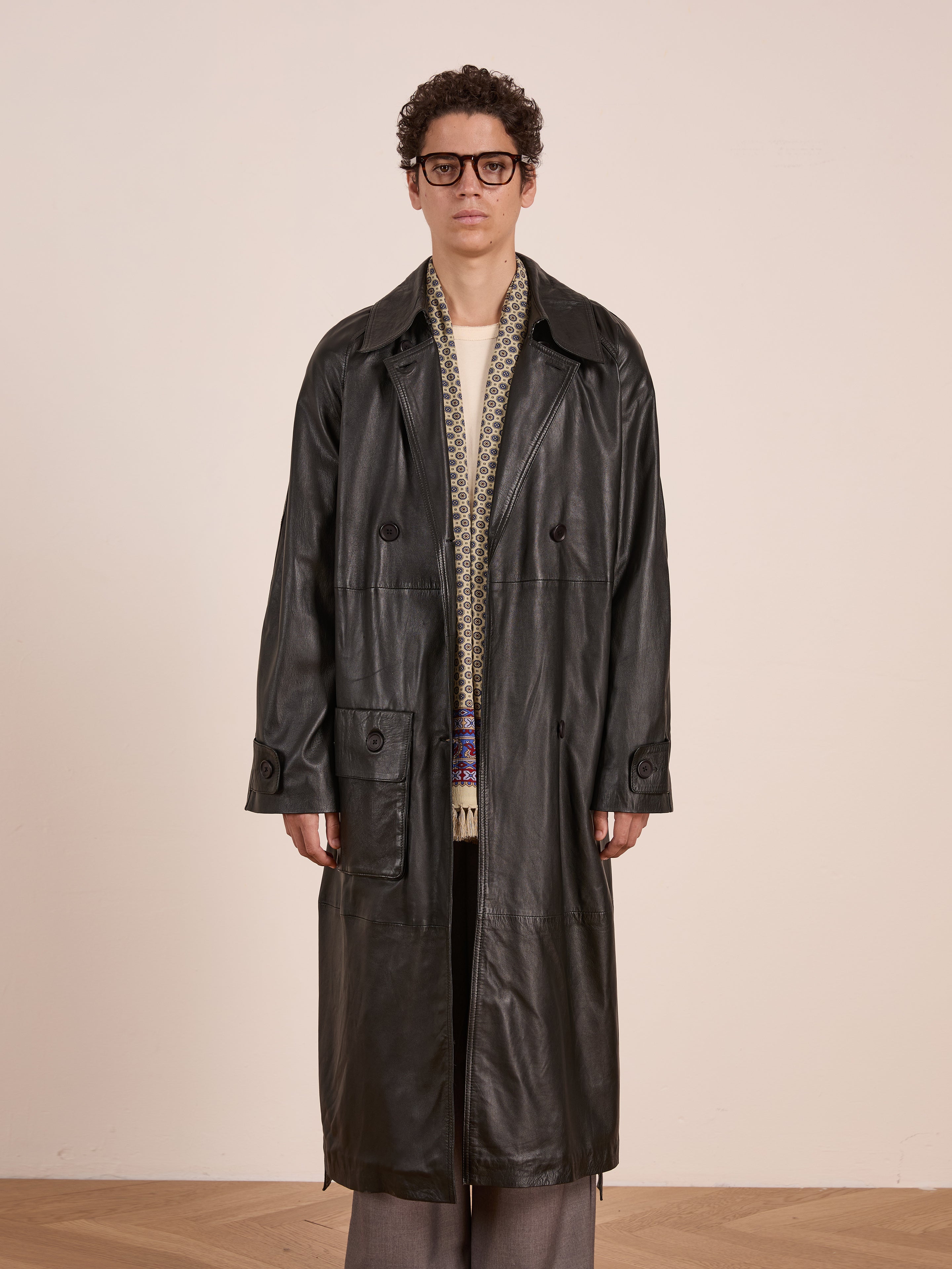 A man with short curly hair and glasses models the FOUND Leather Trench Coat over a cream sweater, patterned scarf, and gray trousers against a plain light background.