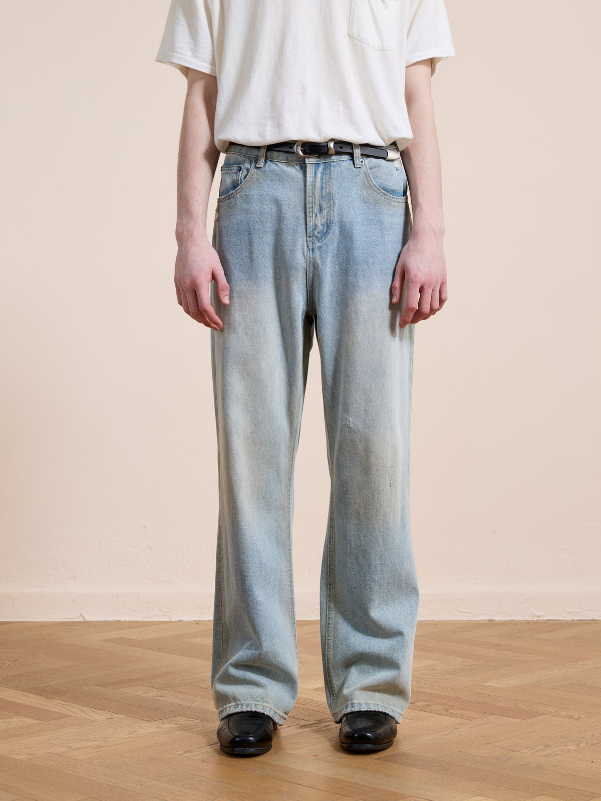 A person stands on a wooden floor against a plain background, wearing Found Straight Leg Jeans in light blue denim, paired with a white t-shirt and black shoes.