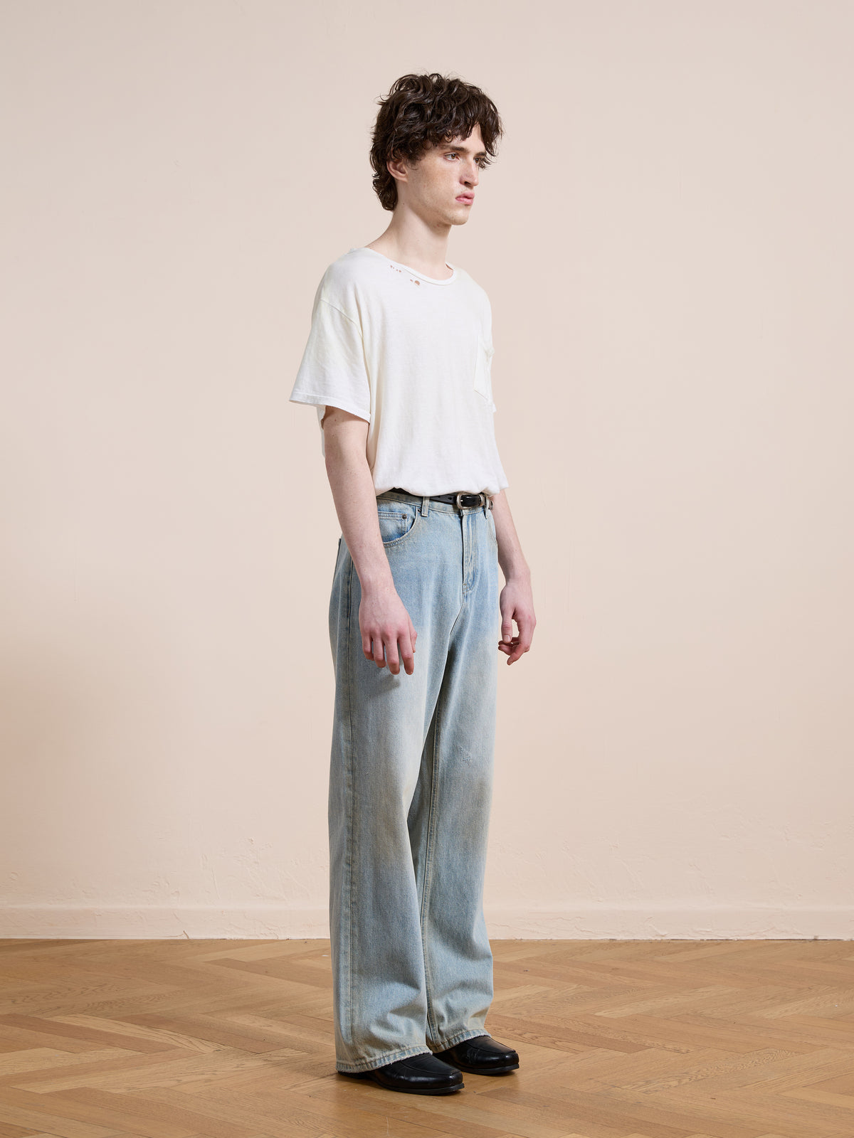 A person stands indoors on a wooden floor, wearing Found Straight Leg Jeans, a white t-shirt, and black shoes while facing slightly to the right against a beige wall.