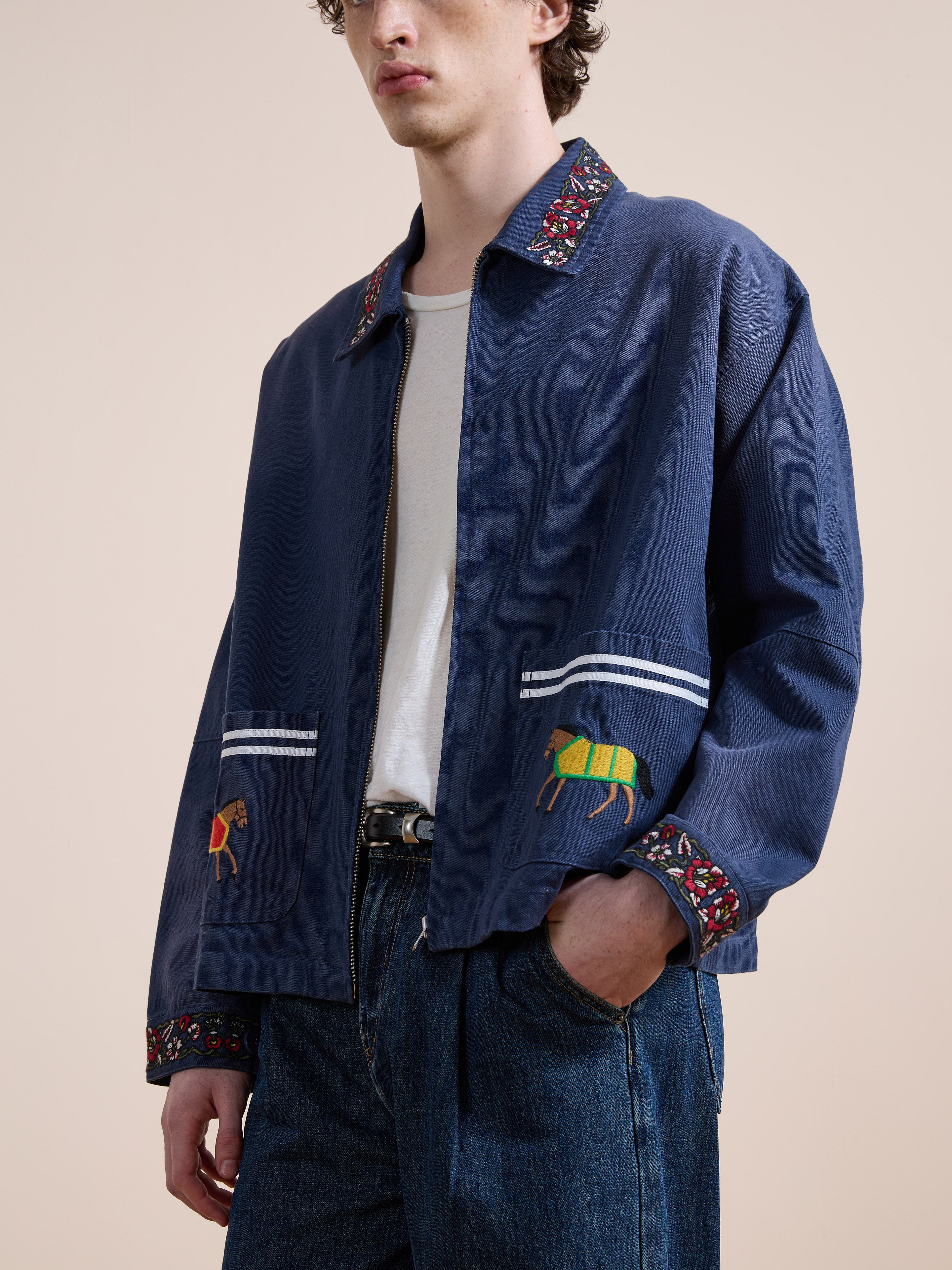A person wears the Found Horse Equine Work Jacket in blue cotton twill, featuring horse embroidery on the pockets and floral collar and cuffs, paired with a white shirt and blue jeans in classic racing colors.