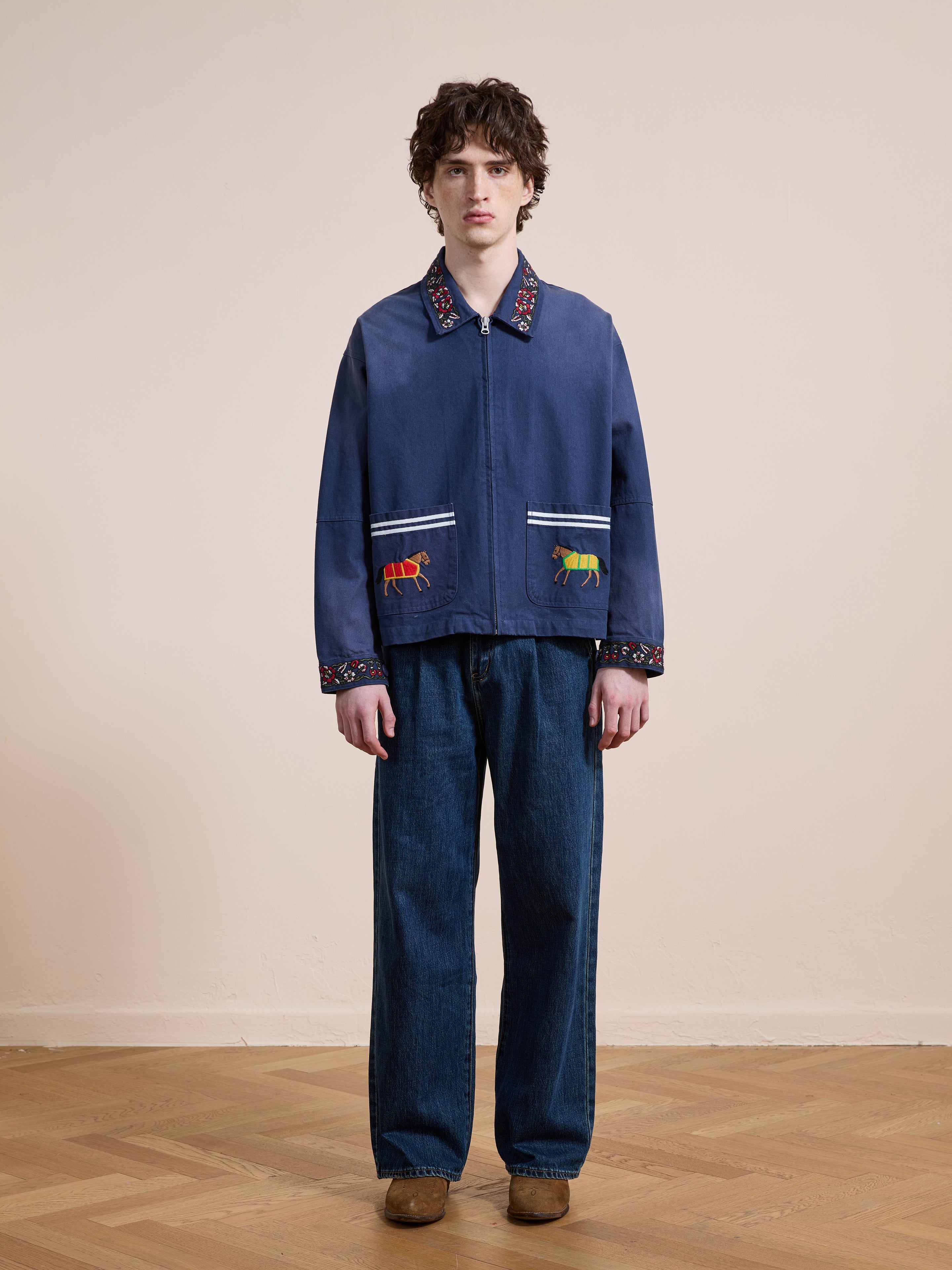 A person stands indoors facing the camera against a plain background, wearing Found’s Horse Equine Work Jacket in cotton twill with colorful embroidery, paired with loose blue jeans and brown shoes.