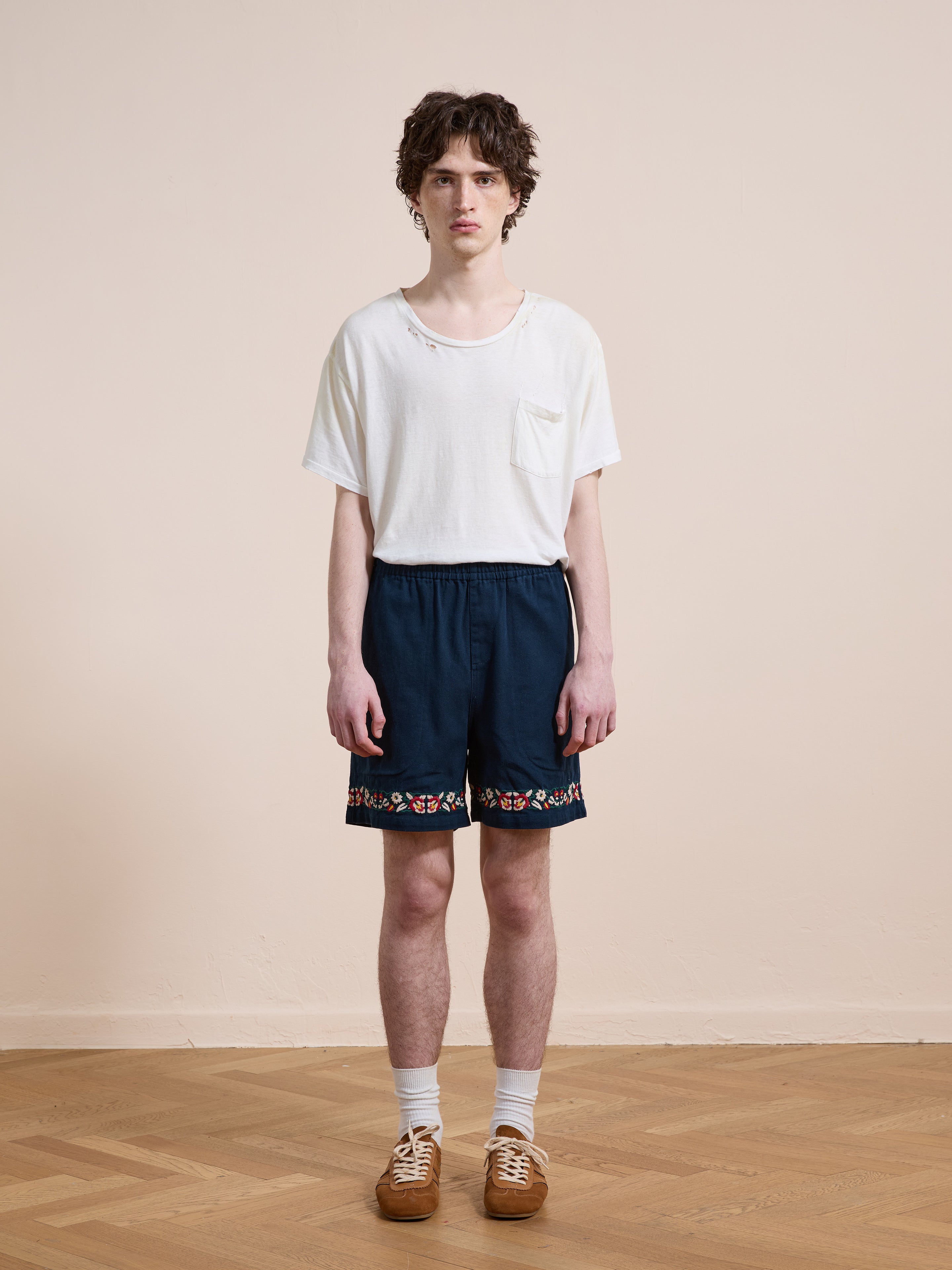 A young person indoors on a wooden floor wears a white t-shirt, Found Horse Equine Twill Shorts with a colorful pattern, white socks, and brown shoes.
