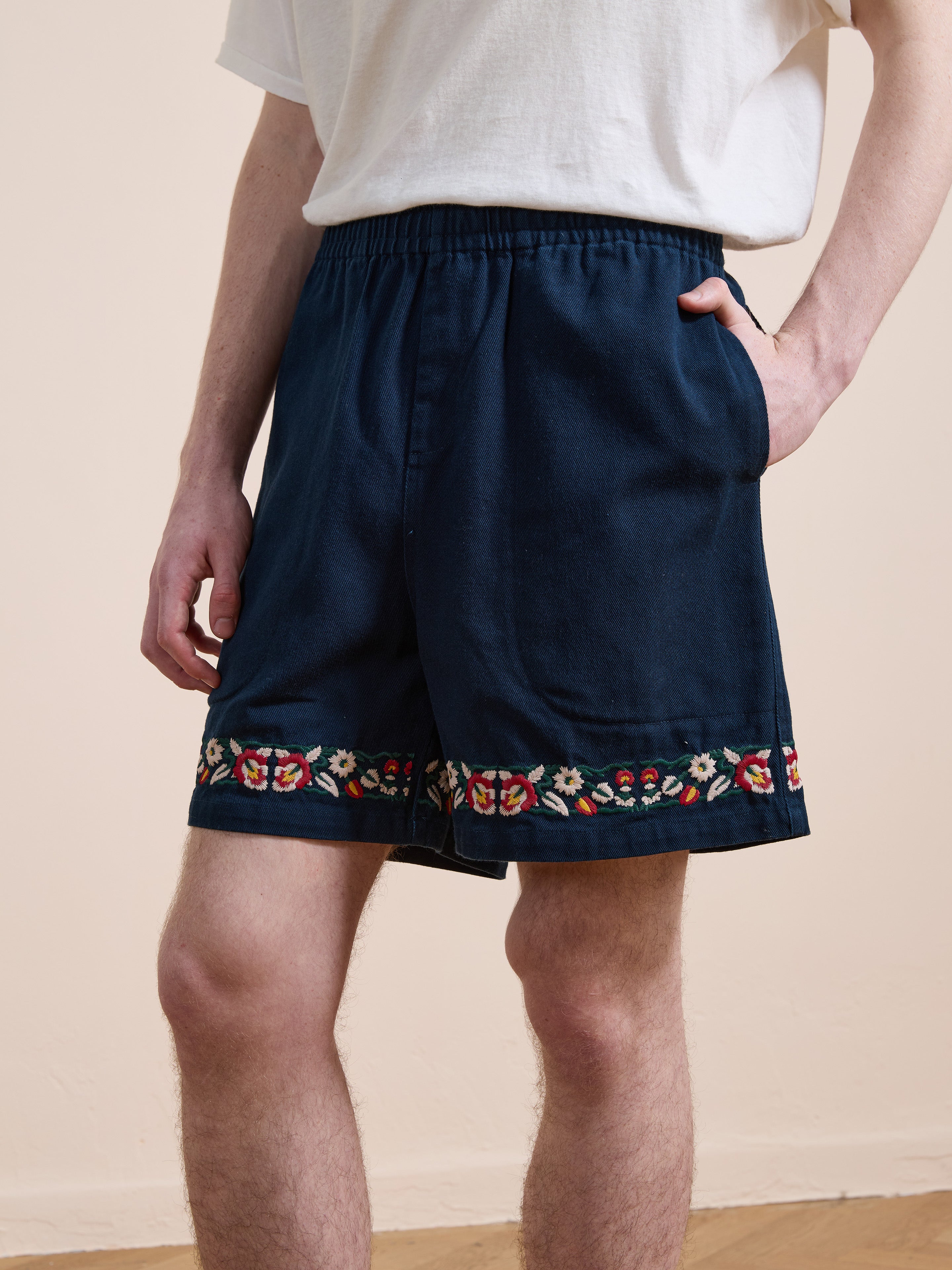 A person wears a white t-shirt and Found Horse Equine Twill Shorts with floral embroidery near the hem, standing indoors with one hand in a pocket.