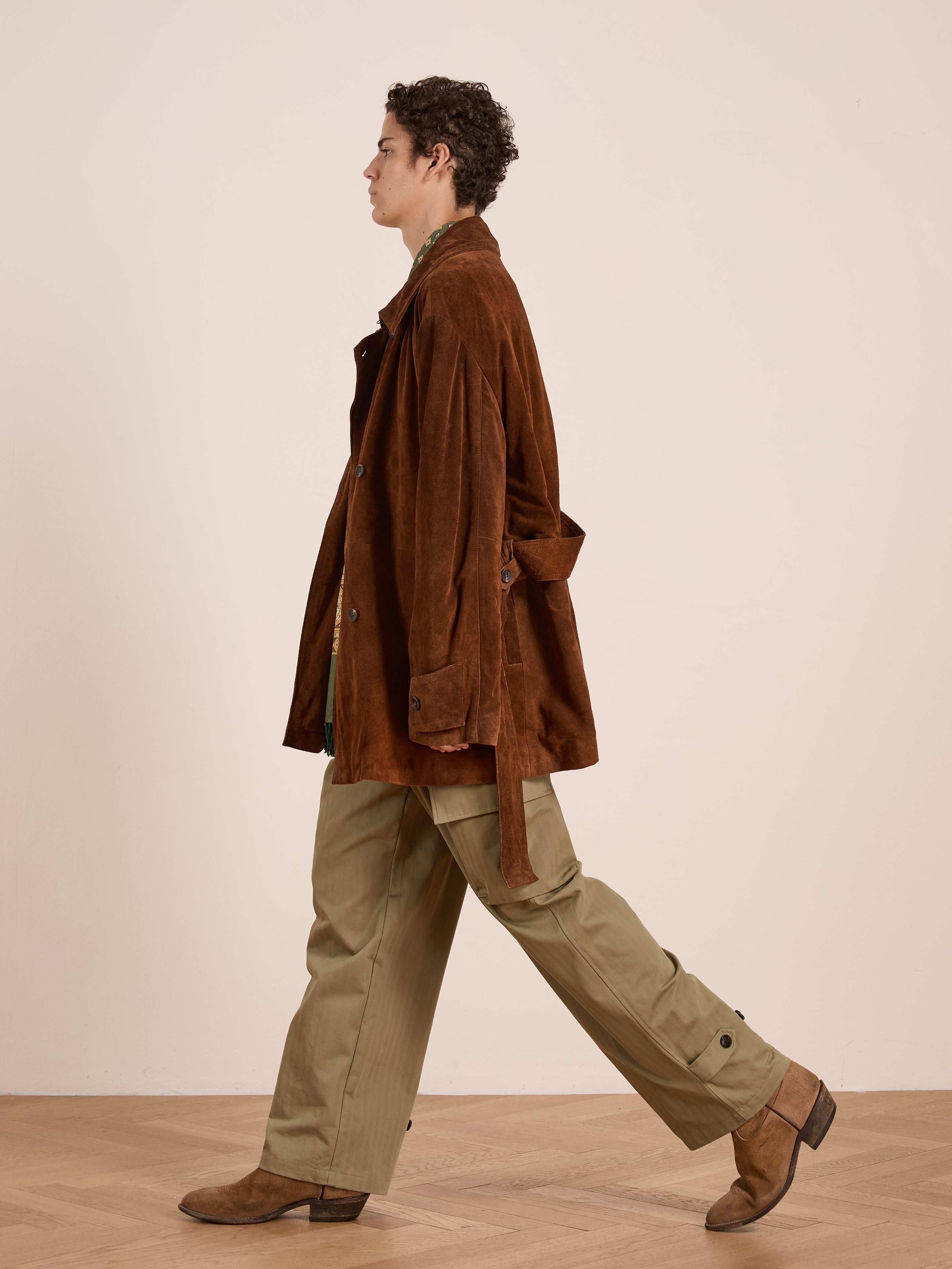 A person walks indoors on a wood floor, wearing a FOUND Double Breasted Suede Leather Jacket, green shirt, khaki pants, and brown boots against a neutral background.