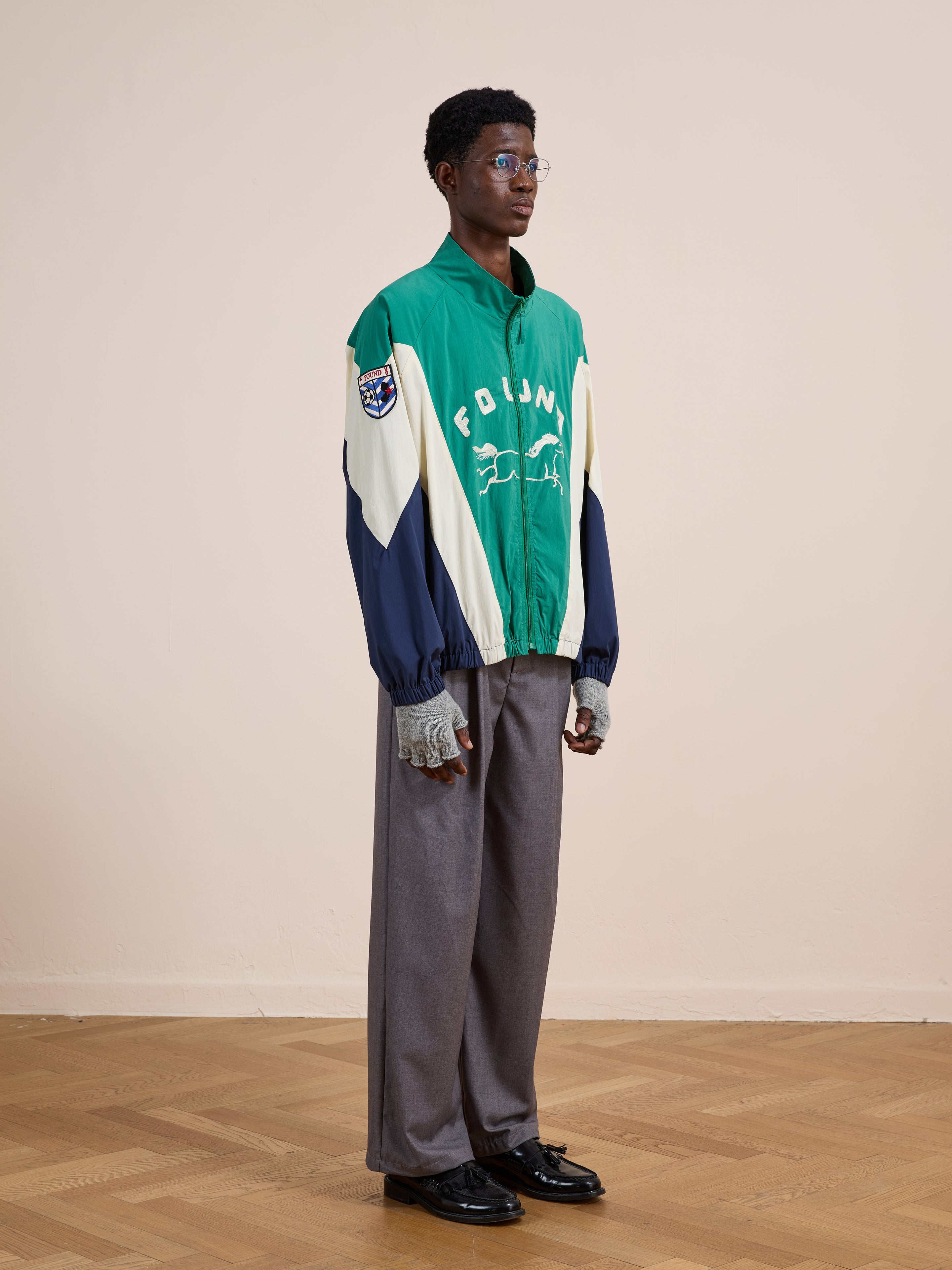 Indoors on a wooden floor, a person wearing glasses, gray pants, black shoes, and a green and cream FOUND Crestline Sport Zip Jacket—a vintage-inspired unisex windbreaker—looks off to the side.