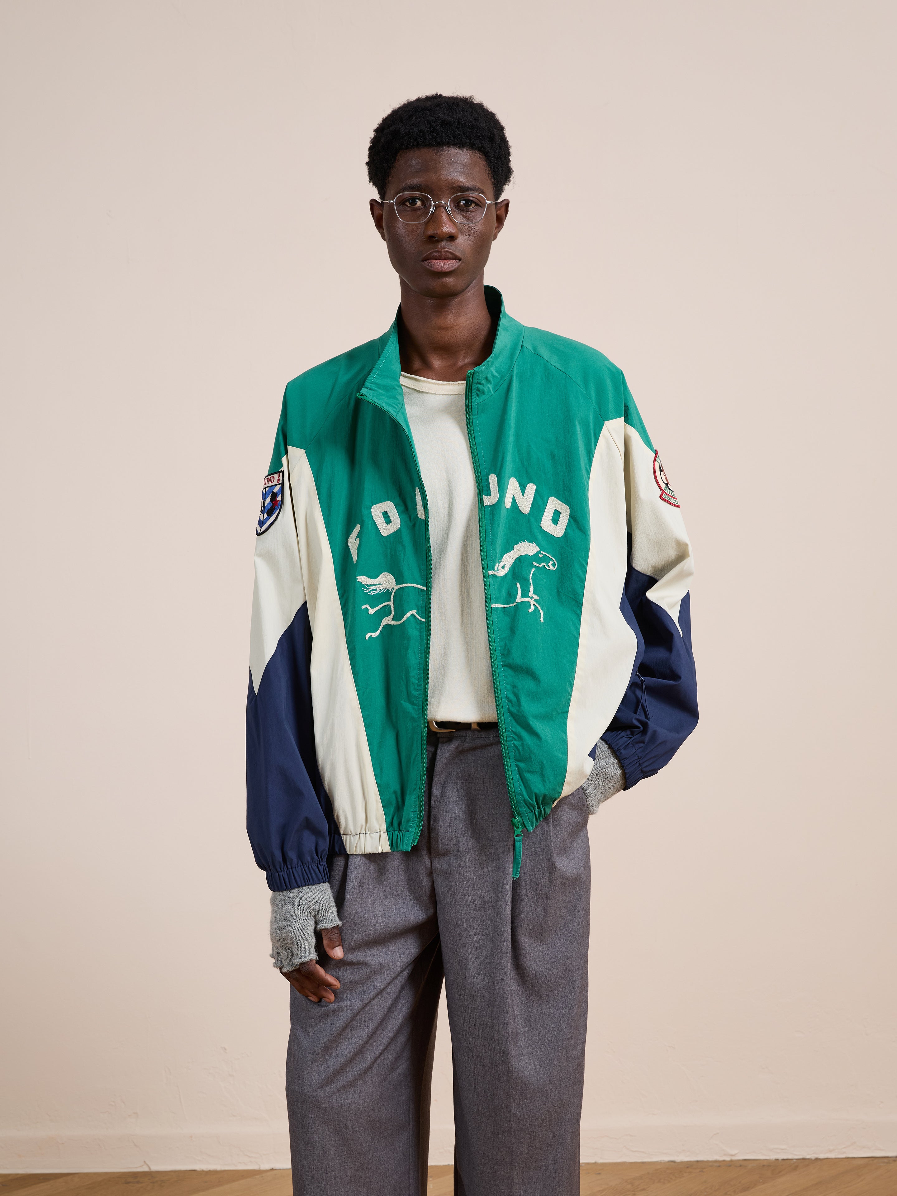 A person wearing glasses, gray trousers, a cream shirt, and the FOUND Crestline Sport Zip Jacket with animal designs stands against a neutral background, highlighting classic vintage sportswear style.
