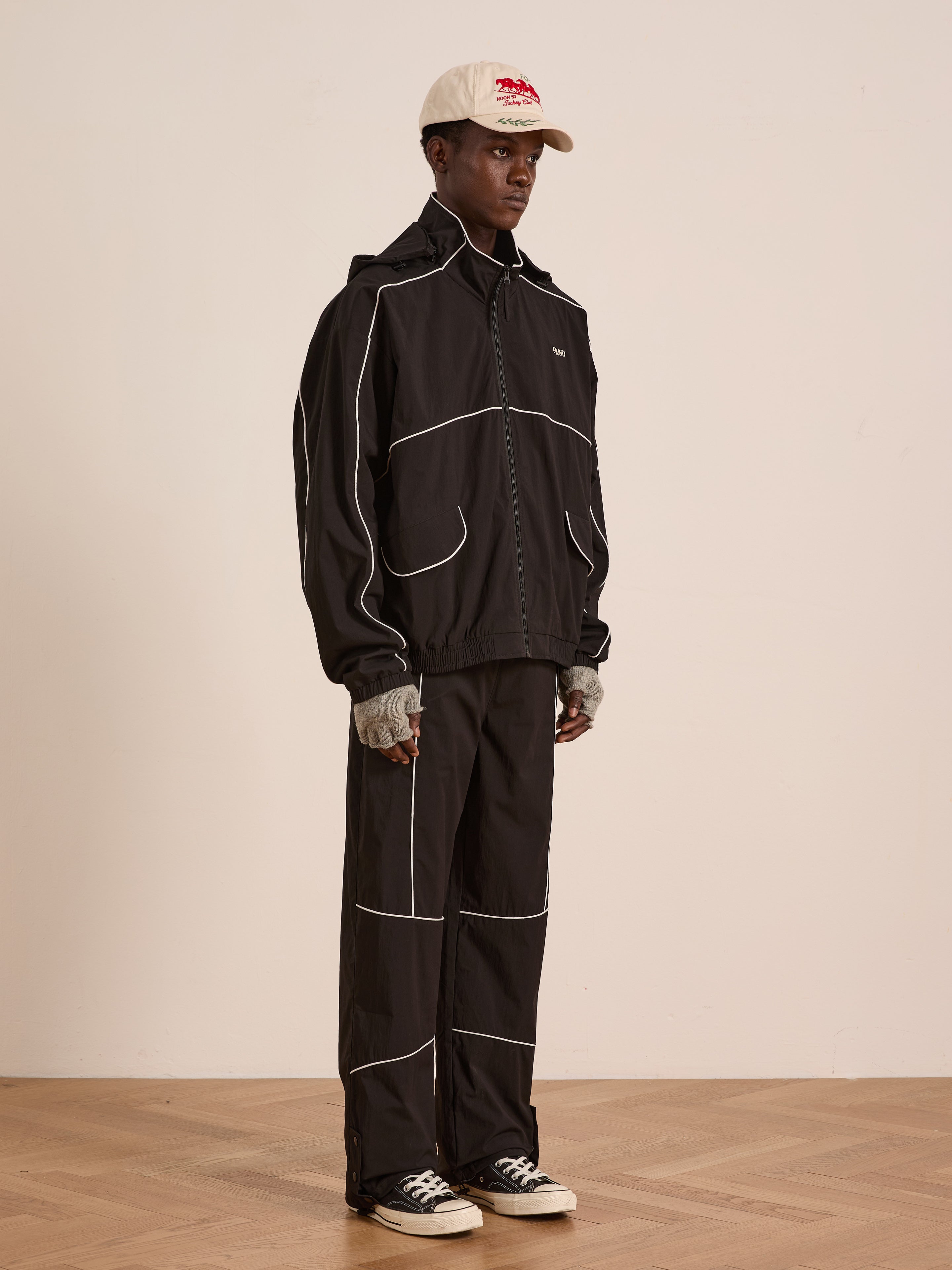 A person in Found Arena Stripe Track Pants—black unisex sportswear with white piping and an adjustable waistband—pairs them with a beige cap, gray fingerless gloves, and black Converse sneakers while standing against a plain wall on a wooden floor.