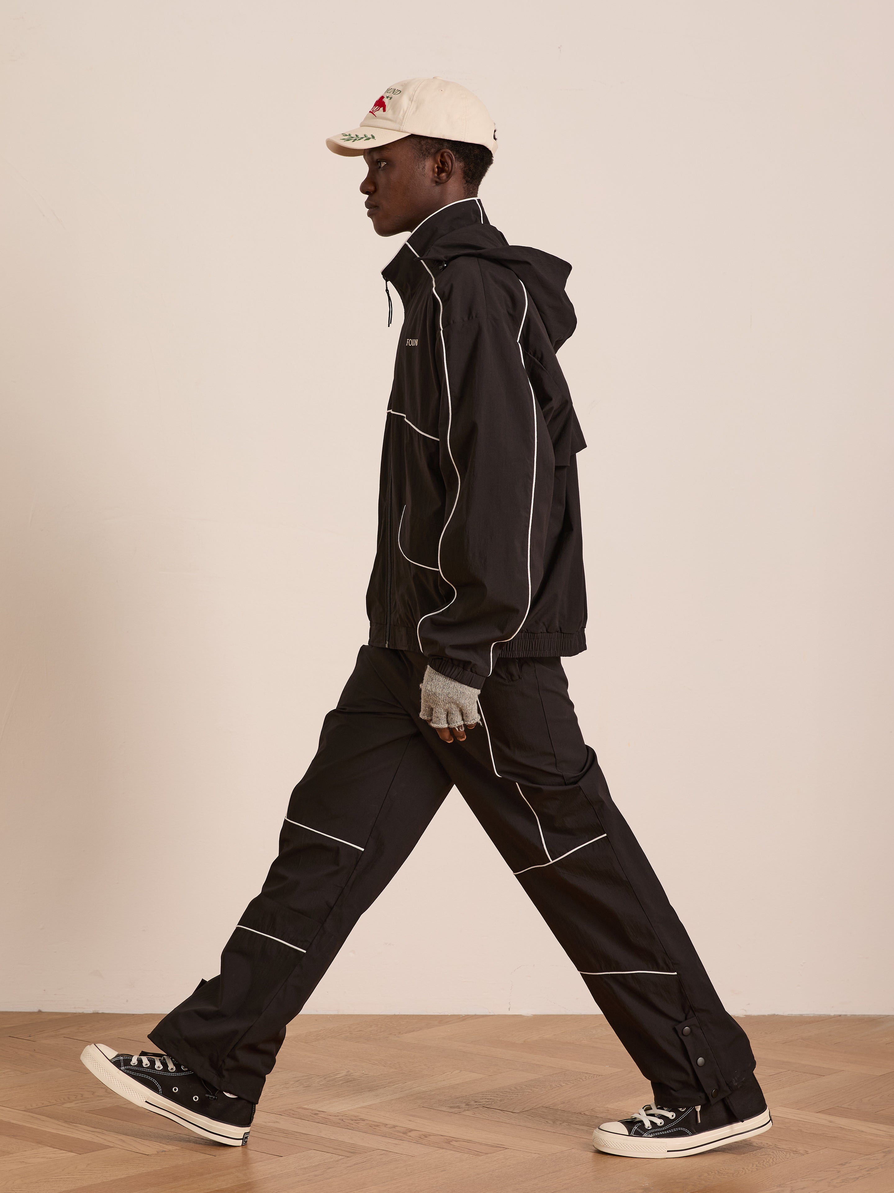 Wearing the FOUND Arena Stripe Track Jacket and matching pants with white piping, a white baseball cap, and black sneakers, a person walks indoors on a wooden floor in style and comfort thanks to the outfit’s lightweight technical fabric.