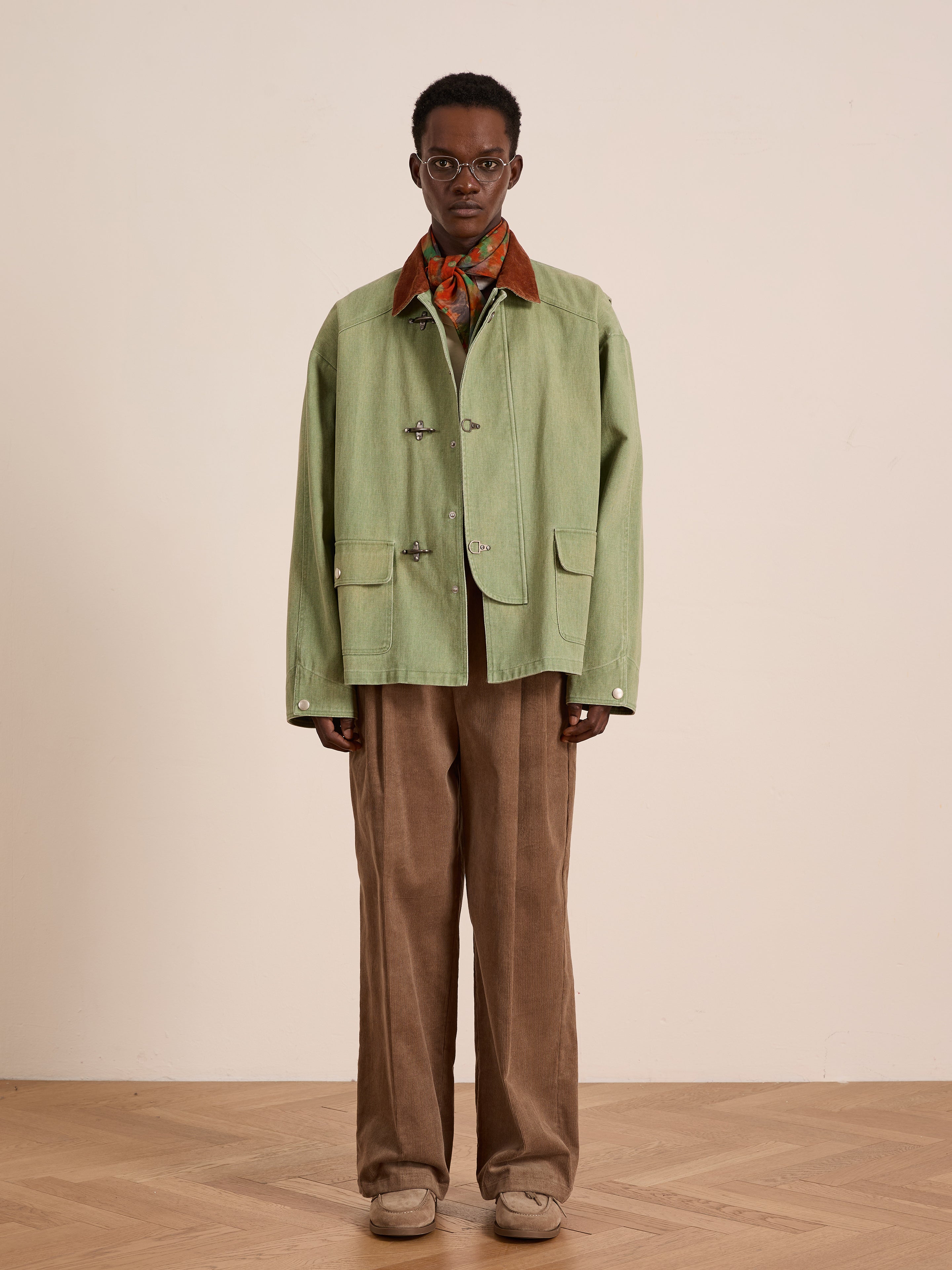 A person stands facing forward wearing glasses and the Found Washed Fireman Clasp Workshop Jacket with a corduroy collar, a patterned scarf, brown wide-leg pants, and beige shoes against a plain background.