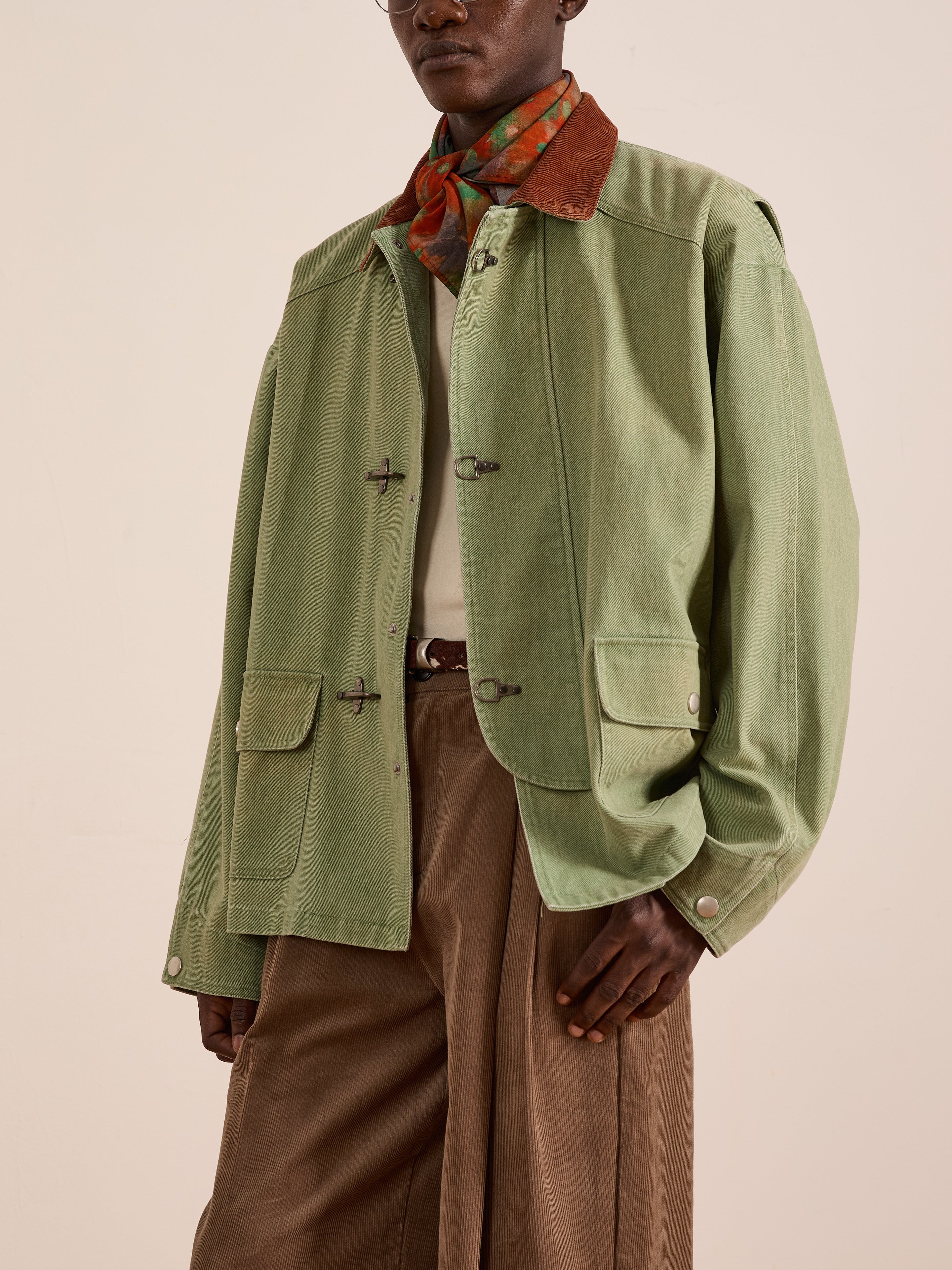 A person wearing Found's Washed Fireman Clasp Workshop Jacket in light green, brown wide-leg pants, a white shirt, and a brown patterned neck scarf stands against a plain beige background.