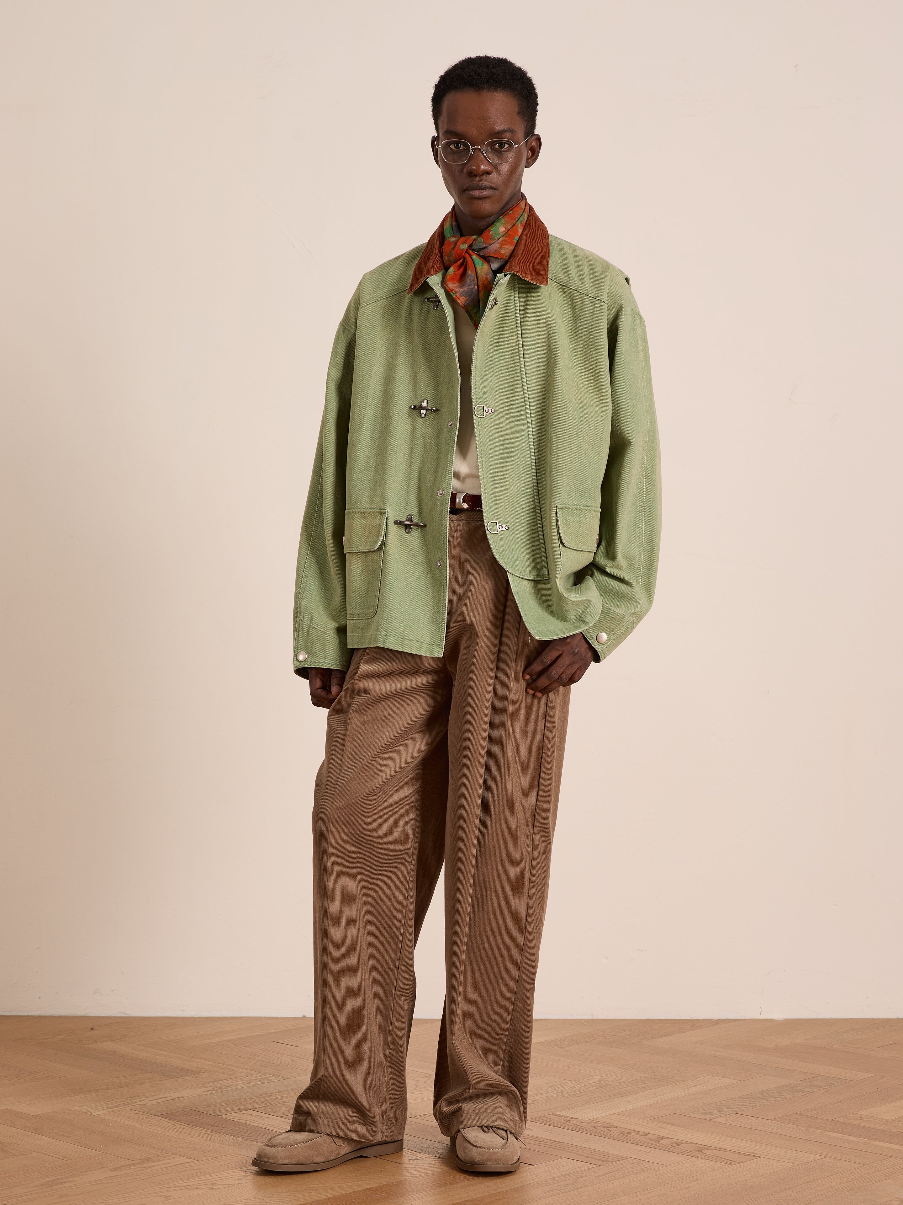 Indoors, a person models Found’s Washed Fireman Clasp Workshop Jacket in light green with a corduroy collar, styled with brown wide-leg pants, tan shoes, a patterned scarf, and glasses against a plain light wall.
