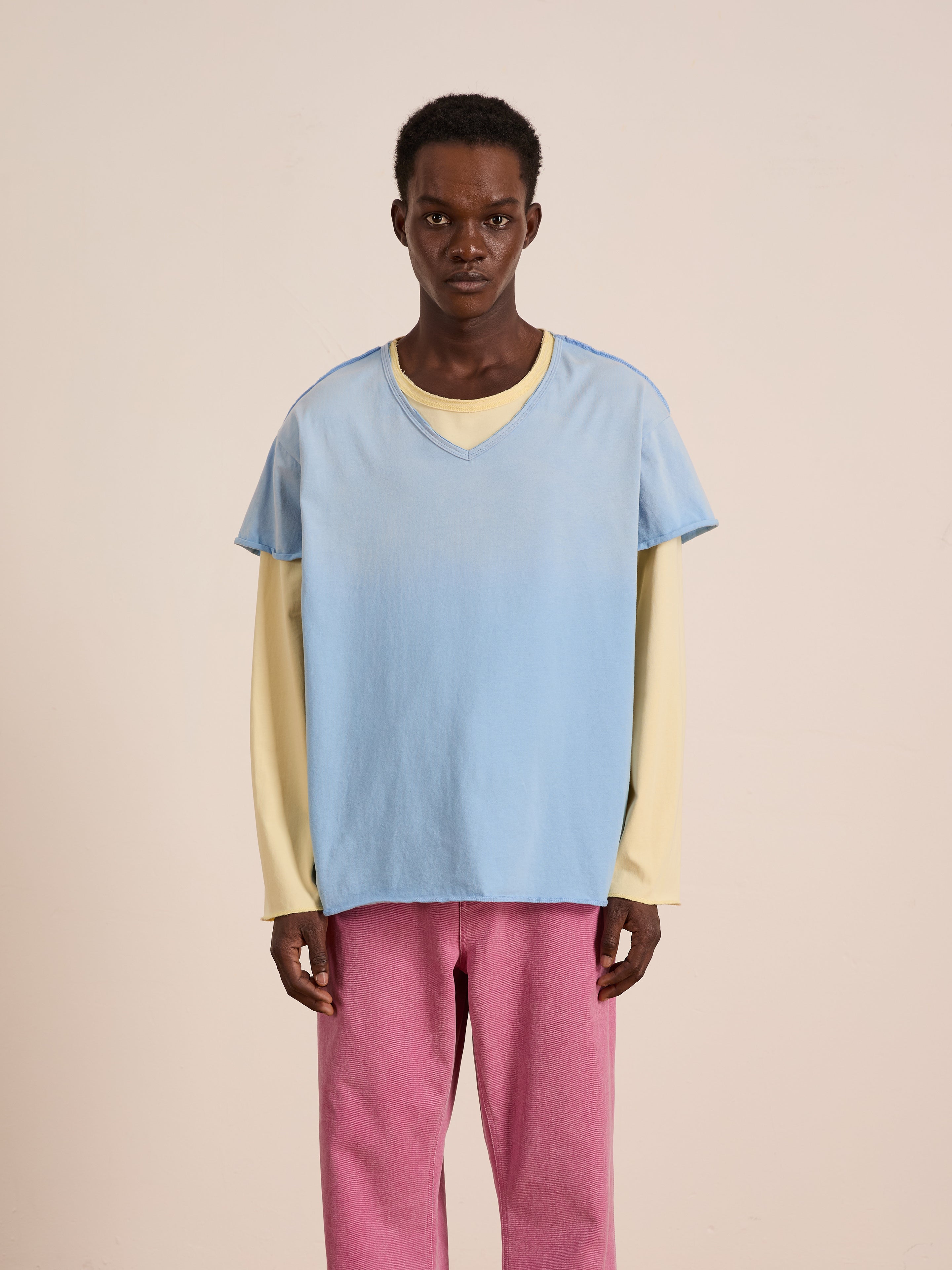 A person faces forward against a plain light background, wearing FOUND's Double Layer LS Tee in pale yellow under a light blue short-sleeve shirt, paired with pink pants.