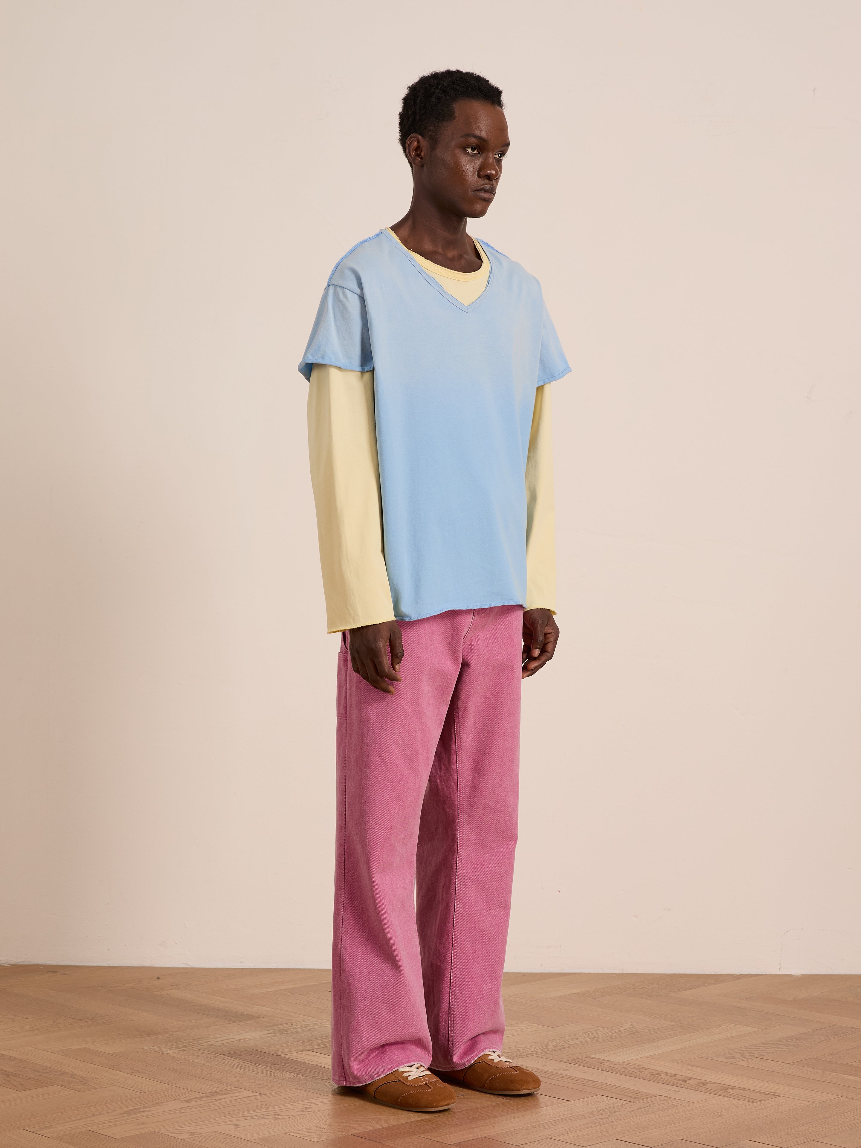 A person indoors wears a FOUND Double Layer LS Tee in pale yellow under a light blue sunfade short-sleeve shirt, paired with pink wide-leg pants and brown shoes, standing against a light wall and wooden floor.
